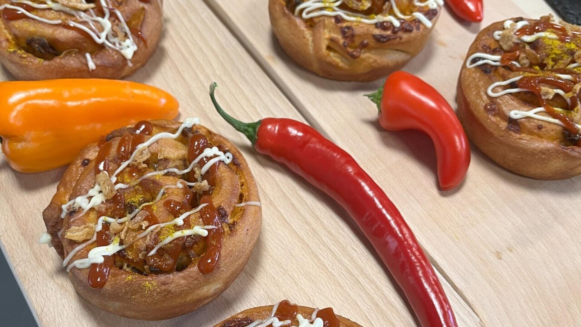 Five savory rolls topped with sauce and drizzled with white dressing are arranged on a wooden board, surrounded by an orange bell pepper and two red chili peppers.