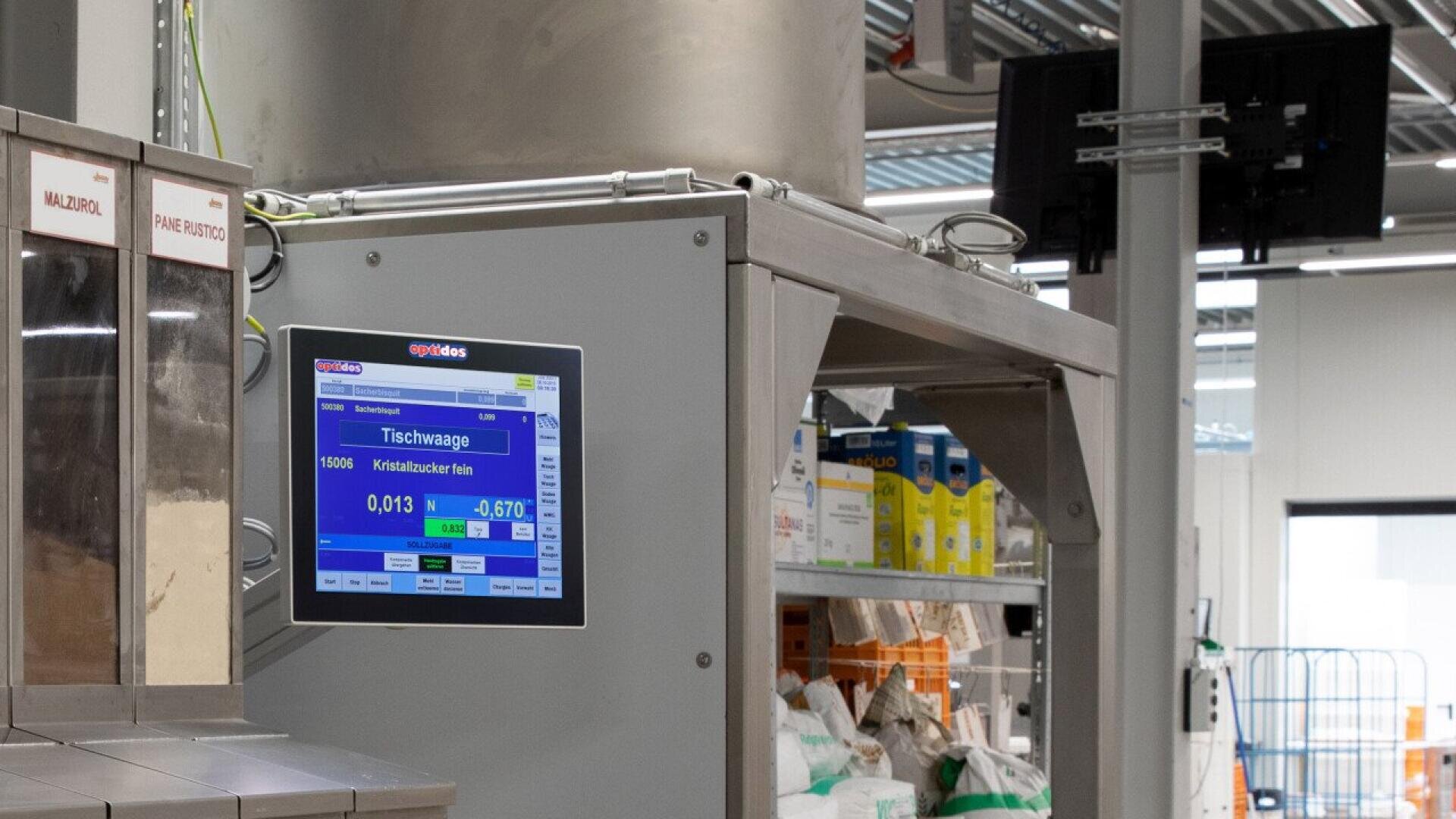 A close-up of an industrial weighing machine with a digital display showing measurements. In the background, shelves with containers and bags are visible in a brightly lit, modern factory or warehouse setting.