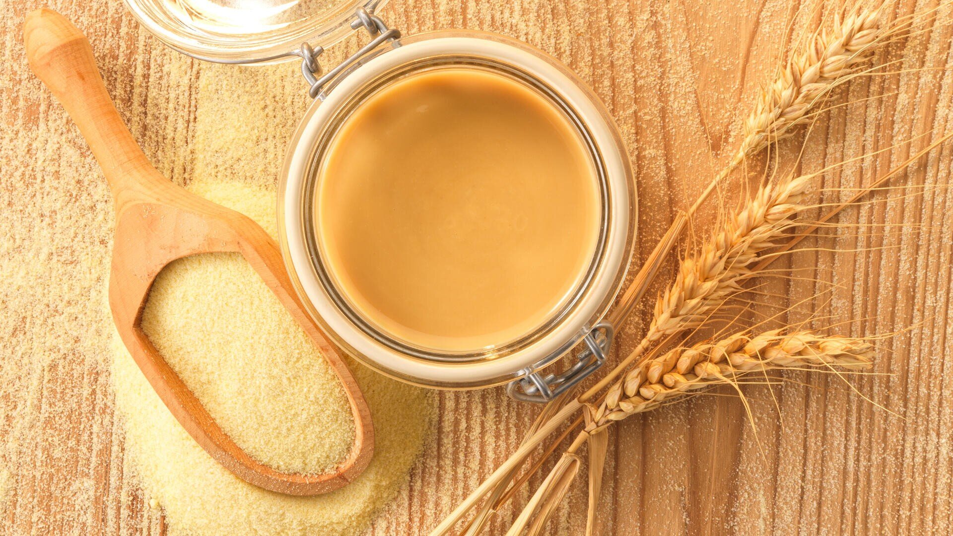 Ein Glas Tahini auf einer Holzoberfläche, daneben eine Holzschaufel gefüllt mit Grieß und einigen Weizenhalmen.