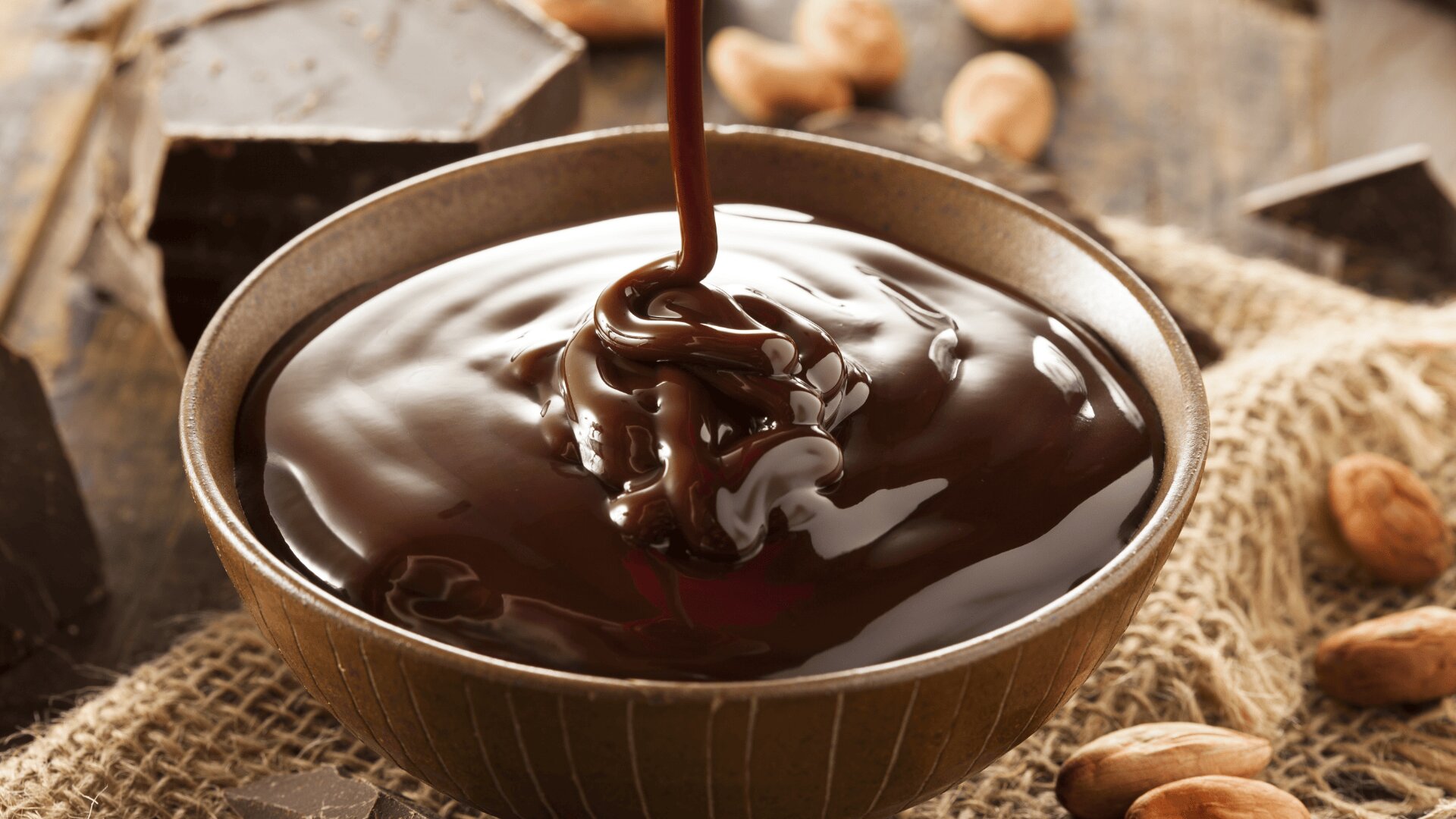A close-up of rich, melted chocolate being poured into a brown bowl, with chocolate chunks and cocoa beans scattered on burlap in the background.