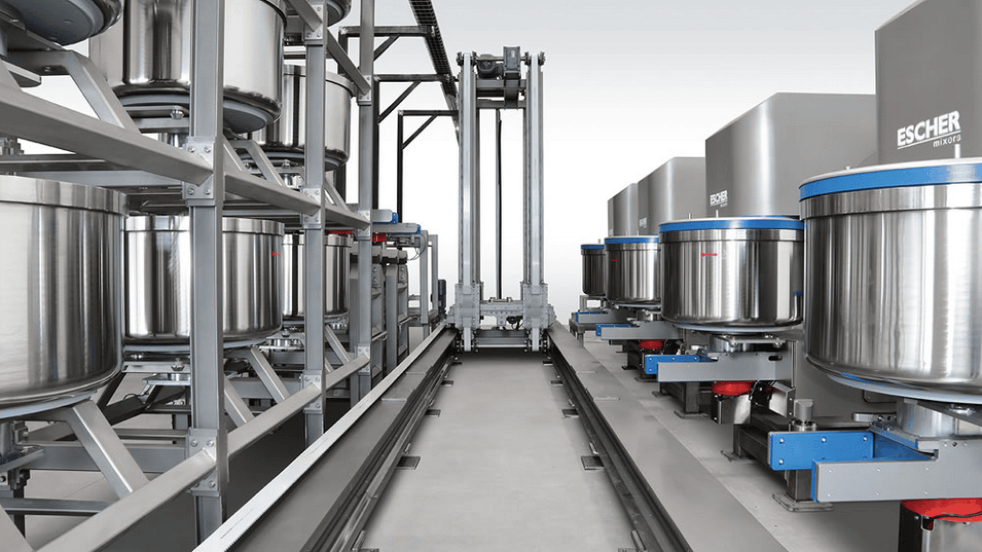 A modern industrial bakery with large stainless steel dough mixers lined up on both sides of a central automated track system for moving mixing bowls.