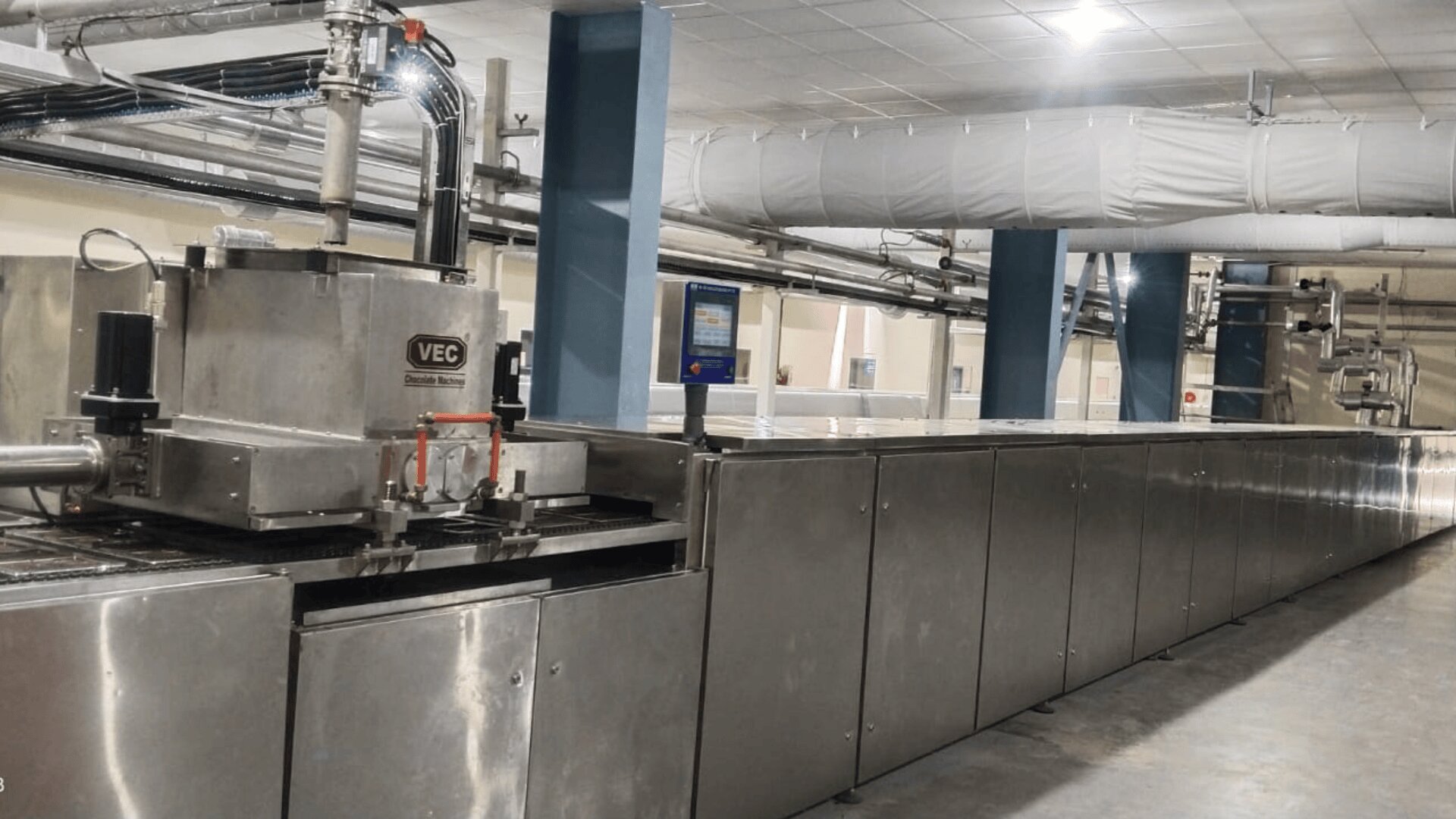 A large, stainless steel industrial food processing machine with control panels and piping in a clean, well-lit factory setting.