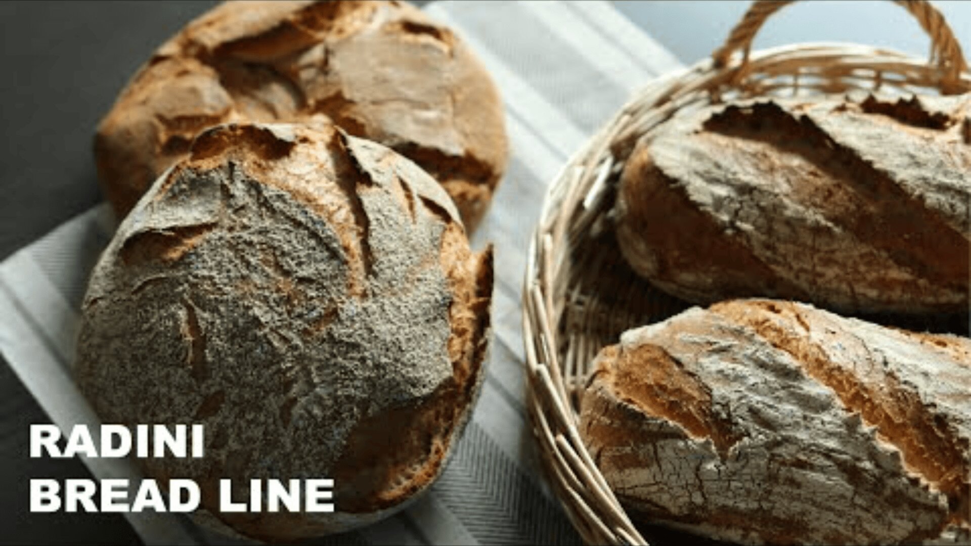 Auf einem gestreiften Tuch und in einem geflochtenen Korb liegen runde und längliche, handwerklich gebackene Brote. Das Brot hat eine knusprige, mehlbestäubte Oberfläche. In der linken unteren Ecke steht der Text "RADINI BREAD LINE".