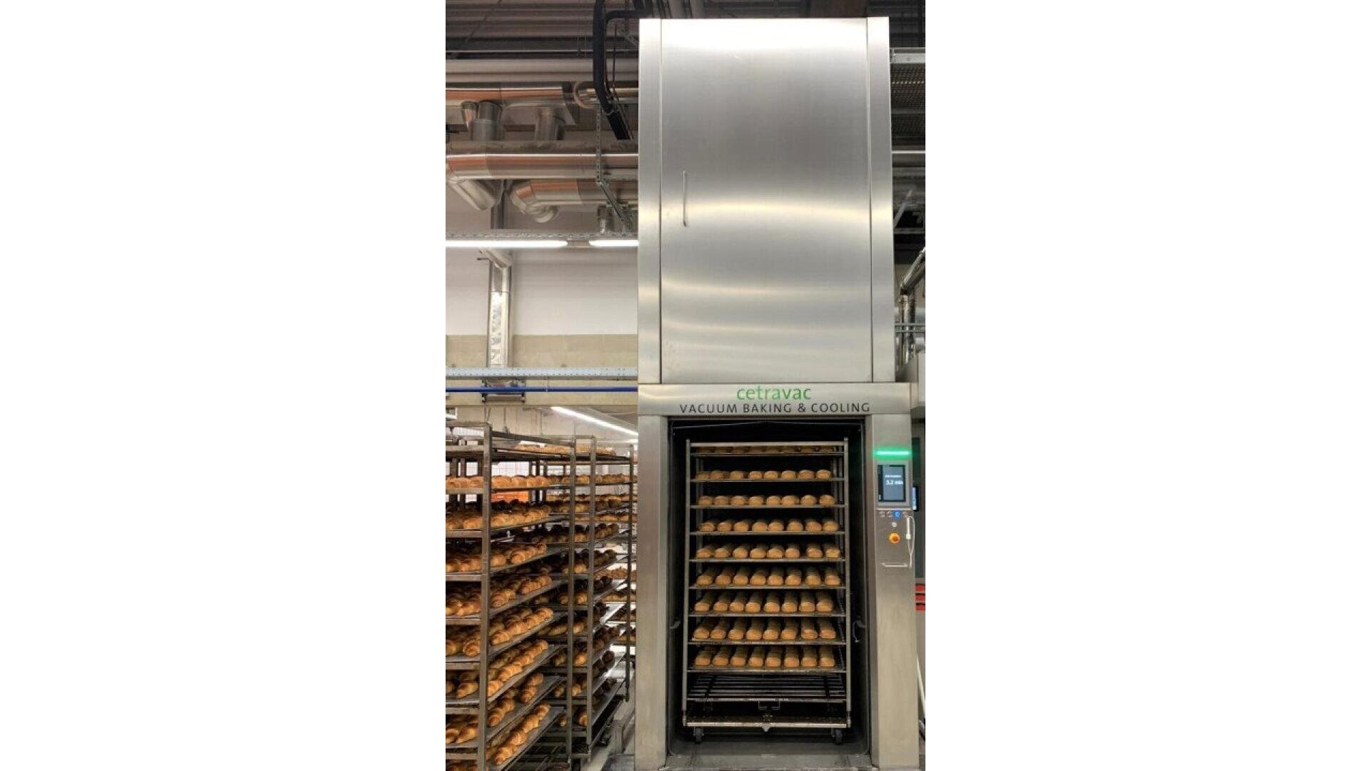 Eine große industrielle Back- und Kühlmaschine mit Tabletts mit gebackenem Brot darin. Auf der linken Seite des Bildes sind Metallregale mit weiteren Brottabletts zu sehen, die in einer Bäckerei oder Fabrik stehen.