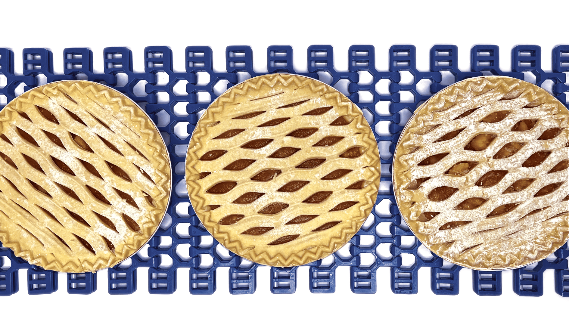 Drei Kuchen mit Gittermuster liegen von oben auf einem blauen Förderband. Die Kuchen haben ein goldenes, kreuz und quer verlaufendes Teigmuster und sind in einer Reihe vor einem weißen Hintergrund angeordnet.