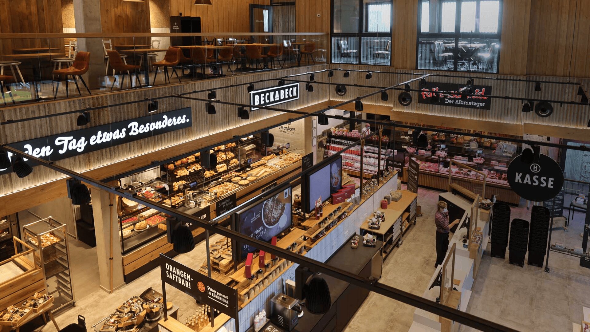 Eine moderne, geräumige Bäckerei und ein Supermarkt mit hölzernen Akzenten, Auslagen für Backwaren, digitalen Speisekarten und Sitzgelegenheiten in einem oberen Zwischengeschoss. In dem gut beleuchteten Innenraum sind nur wenige Kunden und Mitarbeiter zu sehen.