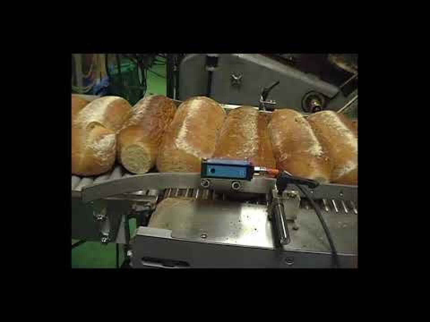 Mehrere mit Mehl bestäubte Brotlaibe laufen in einer Fabrik oder Industrieanlage über ein Förderband, an dem ein Überwachungsgerät angebracht ist.