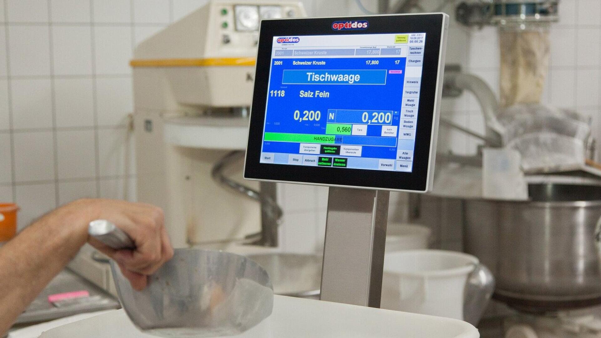 A person uses a metal scoop near an electronic scale with a display showing measurements in grams in a commercial kitchen or bakery setting. White tiled walls and baking equipment are visible in the background.