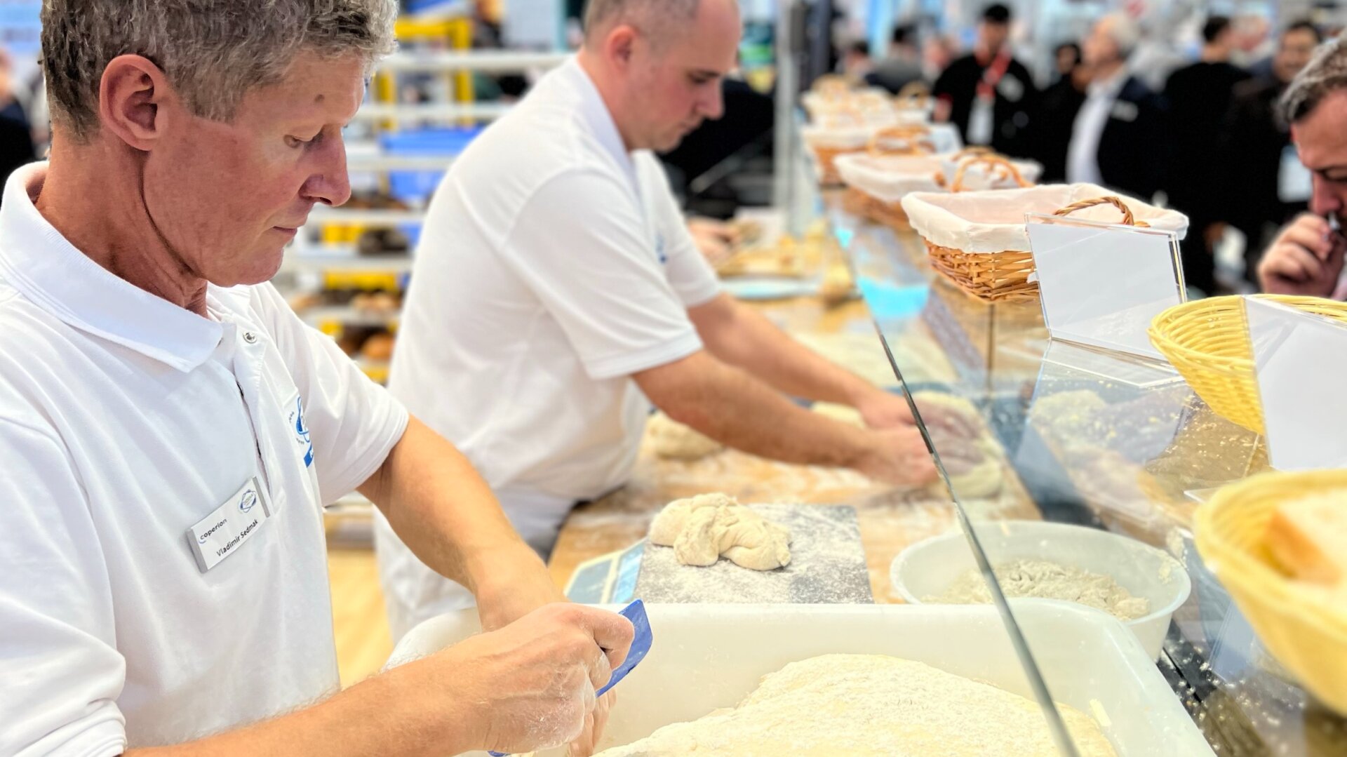 Zwei Männer in weißen Uniformen bereiten an einer belebten Theke in einer hell erleuchteten Ausstellungshalle Teig zu. Um sie herum sind Backzutaten und Geräte sichtbar. Im Hintergrund sind Menschen und Schilder zu sehen.