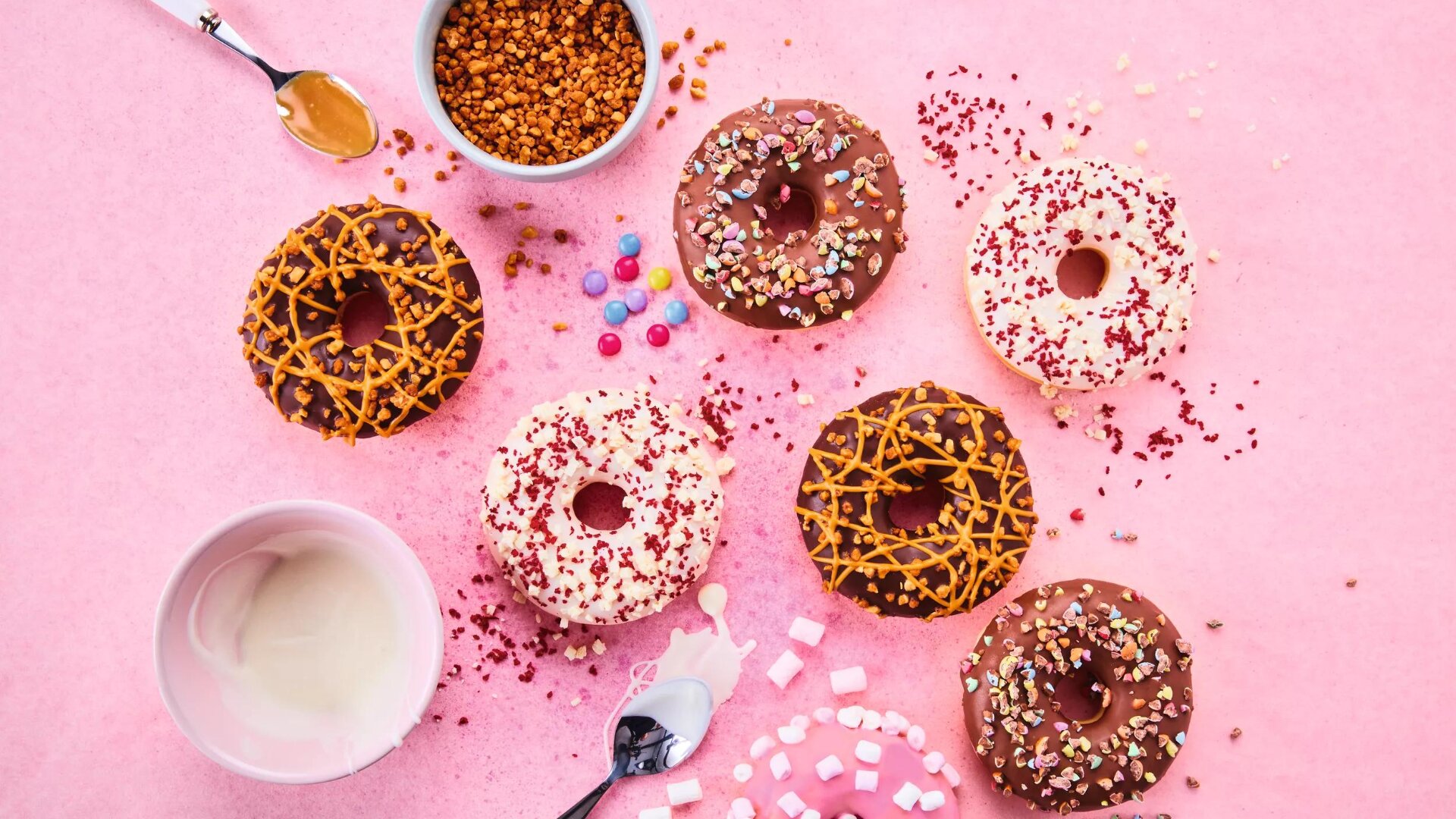 Bunte Donuts mit Streuseln und Nieselregen sind auf einer rosafarbenen Fläche arrangiert, umgeben von kleinen Schalen mit Toppings, einem Löffel mit Karamell, verstreuten Marshmallows und bunten Bonbons.