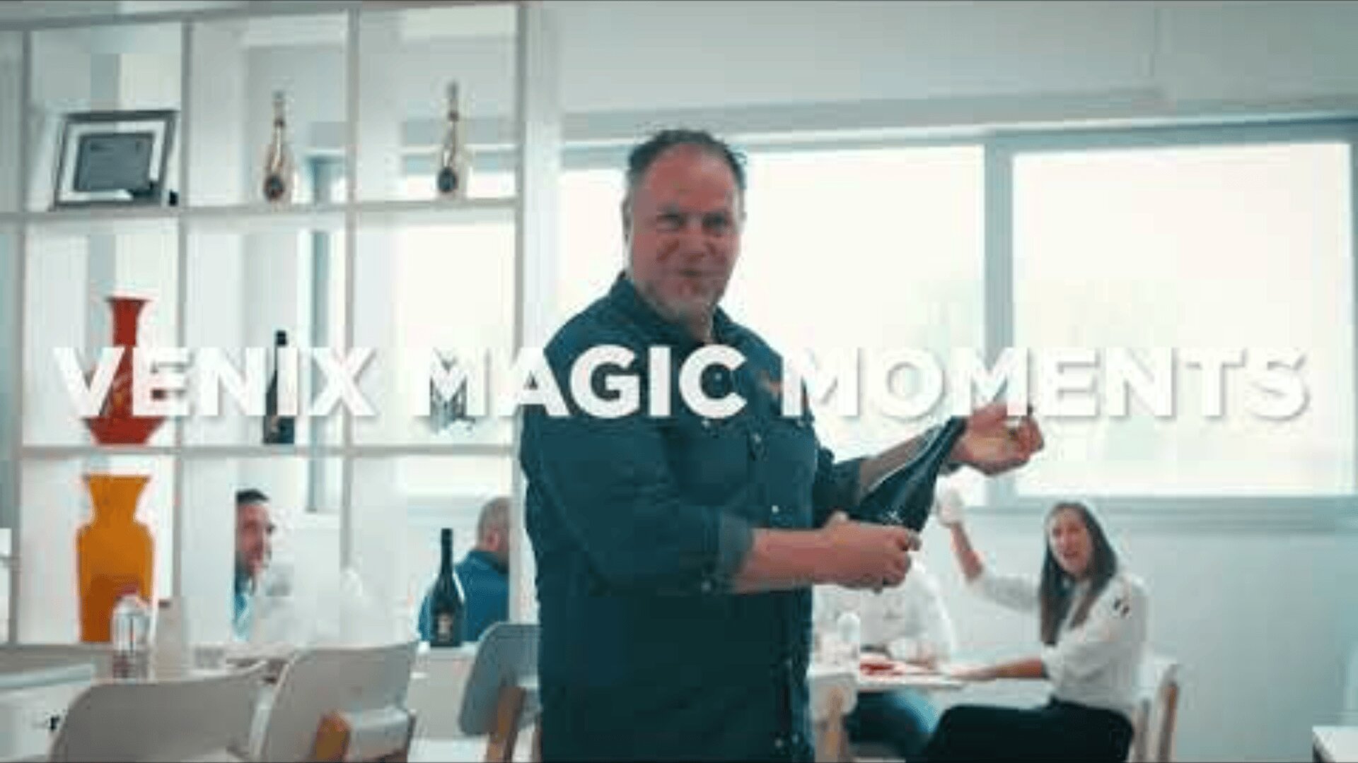 A man smiles while opening a bottle in a bright room with people sitting at a table in the background. Large text across the image reads “VENIX MAGIC MOMENTS.” Shelves with bottles and vases are visible on the left.