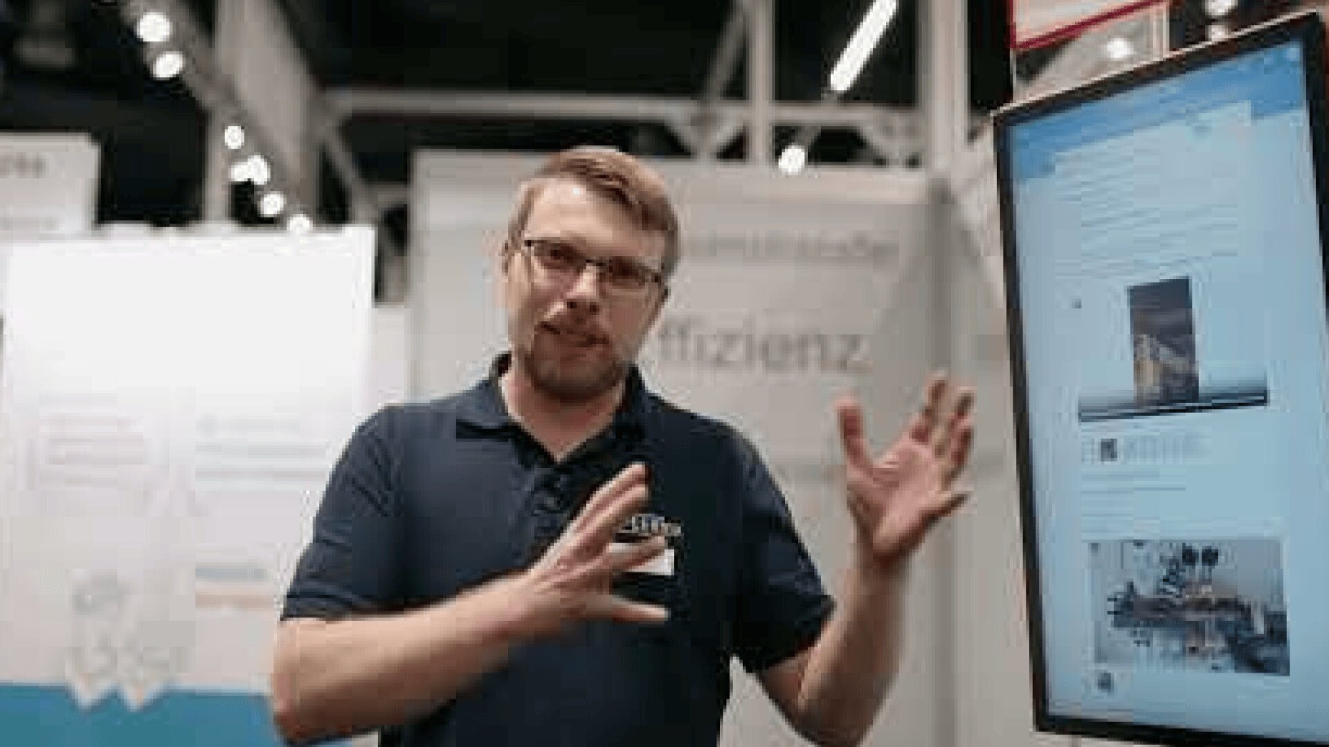 Ein Mann mit Brille und marineblauem Poloshirt gestikuliert mit den Händen, während er neben einem großen vertikalen Monitor steht, auf dem die Website einer scheinbaren Technologie- oder Wirtschaftsausstellung angezeigt wird.