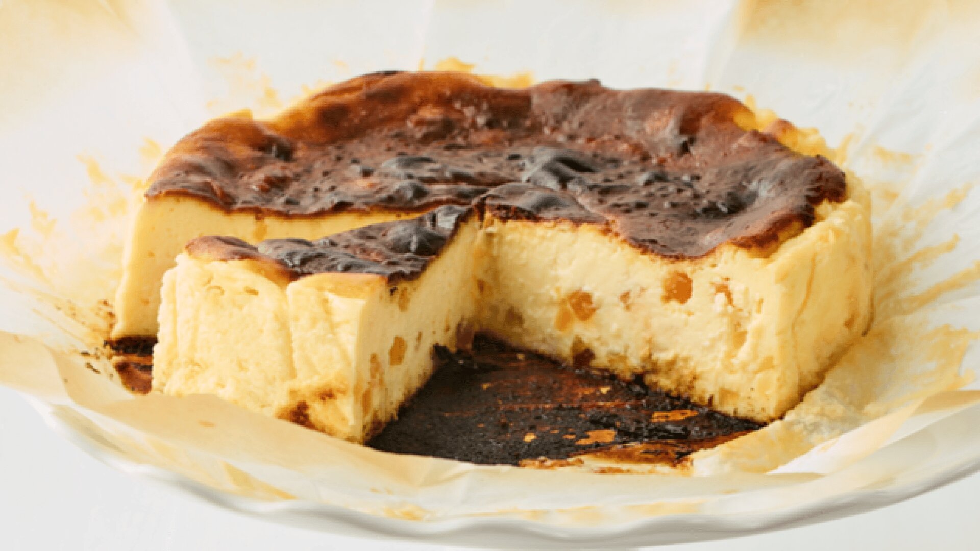 A partially sliced burnt Basque cheesecake on a parchment-lined cake stand, with two slices served on separate plates, each with a fork.