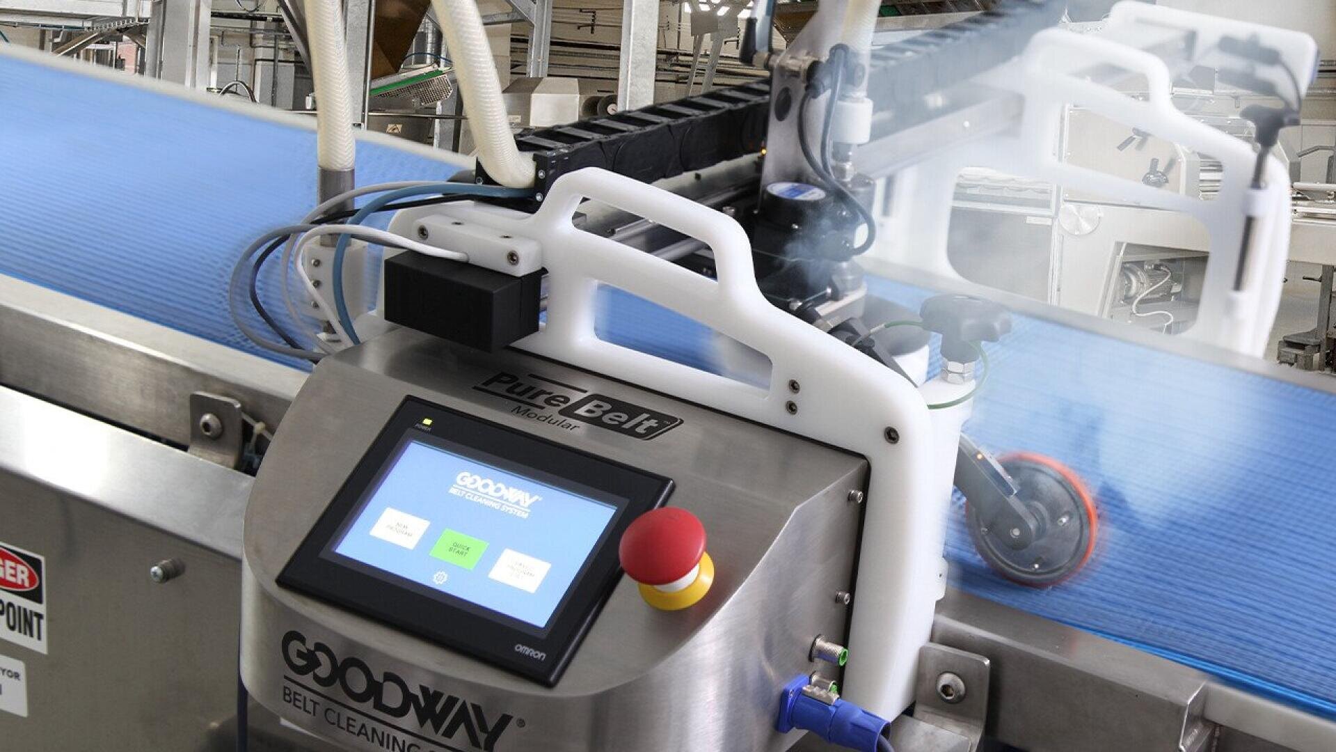 Eine Nahaufnahme eines automatischen Bandreinigungssystems mit einem Bedienfeld an einem Förderband in einer Industrieumgebung. Das System sprüht während der Reinigung des blauen Bandes Nebel.