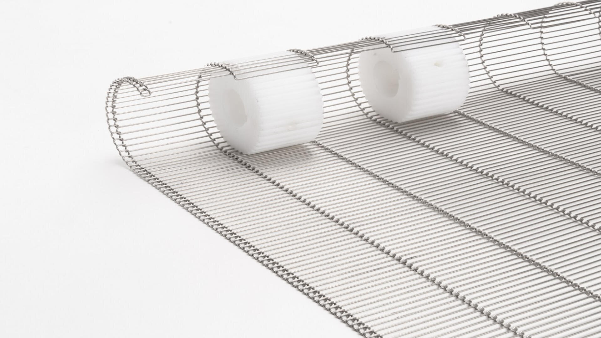 Close-up of a metal wire conveyor belt with evenly spaced, white plastic rollers attached along one edge, placed on a plain white background.