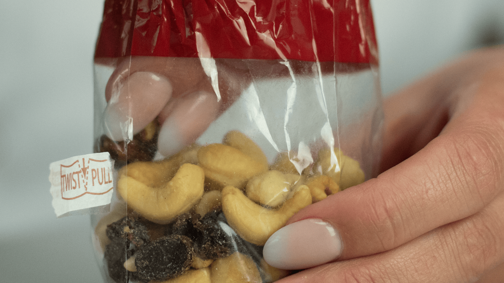 A close-up of hands holding a plastic bag of mixed nuts and raisins, sealed with a red twist tie. The bag has a label that says Twist & Pull. The persons nails are painted with a French manicure.