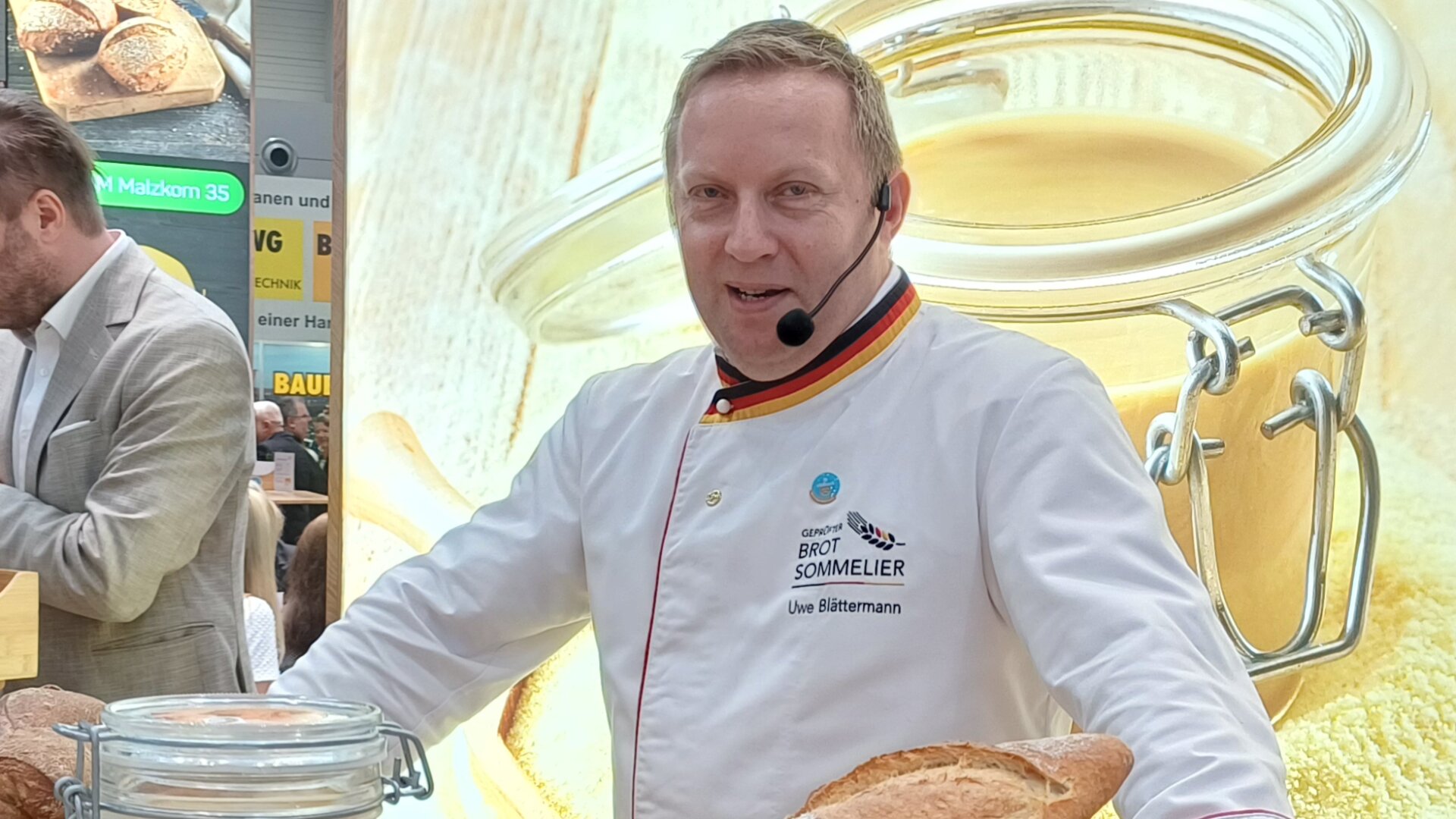 A baker stands behind a table displaying breads, pastries, a large jar of creamy spread, and jars of preserves at a food event. A sign and award are visible, with people interacting in the background.