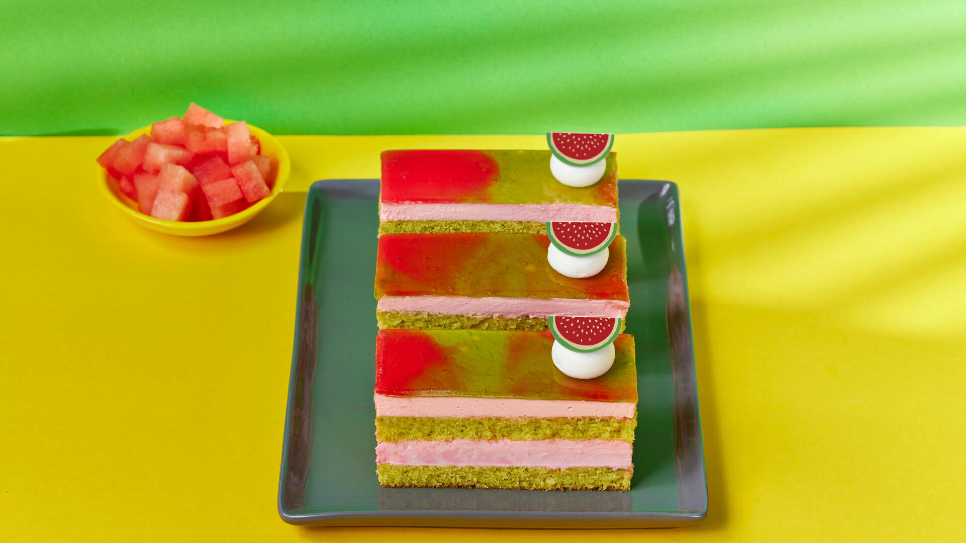 Drei Stücke Schichtkuchen mit Wassermelonenmotiv auf einem grauen Teller, jedes mit dekorativen Wassermelonenstücken belegt, im Hintergrund auf einer gelb-grünen Fläche eine kleine Schale mit Wassermelonenwürfeln.