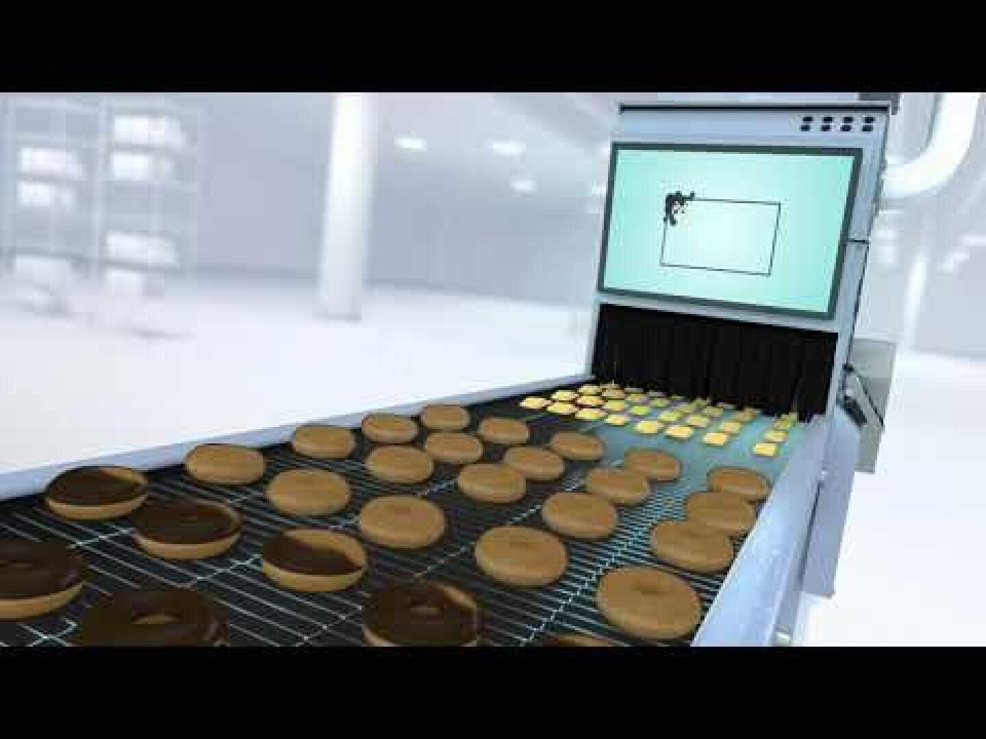 A conveyor belt carries rows of donuts under a machine with a screen displaying a donut image, suggesting an automated quality control or inspection process in a factory setting.