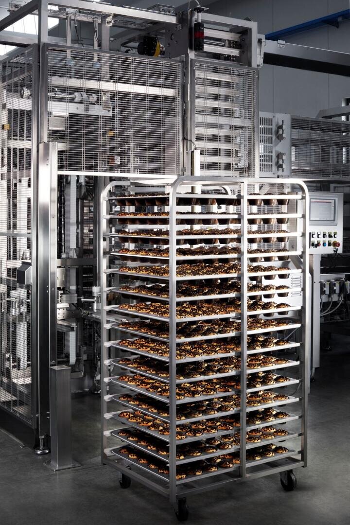 A metal rack with multiple shelves holds trays filled with baked goods, situated in front of large industrial machinery inside a modern food production facility.