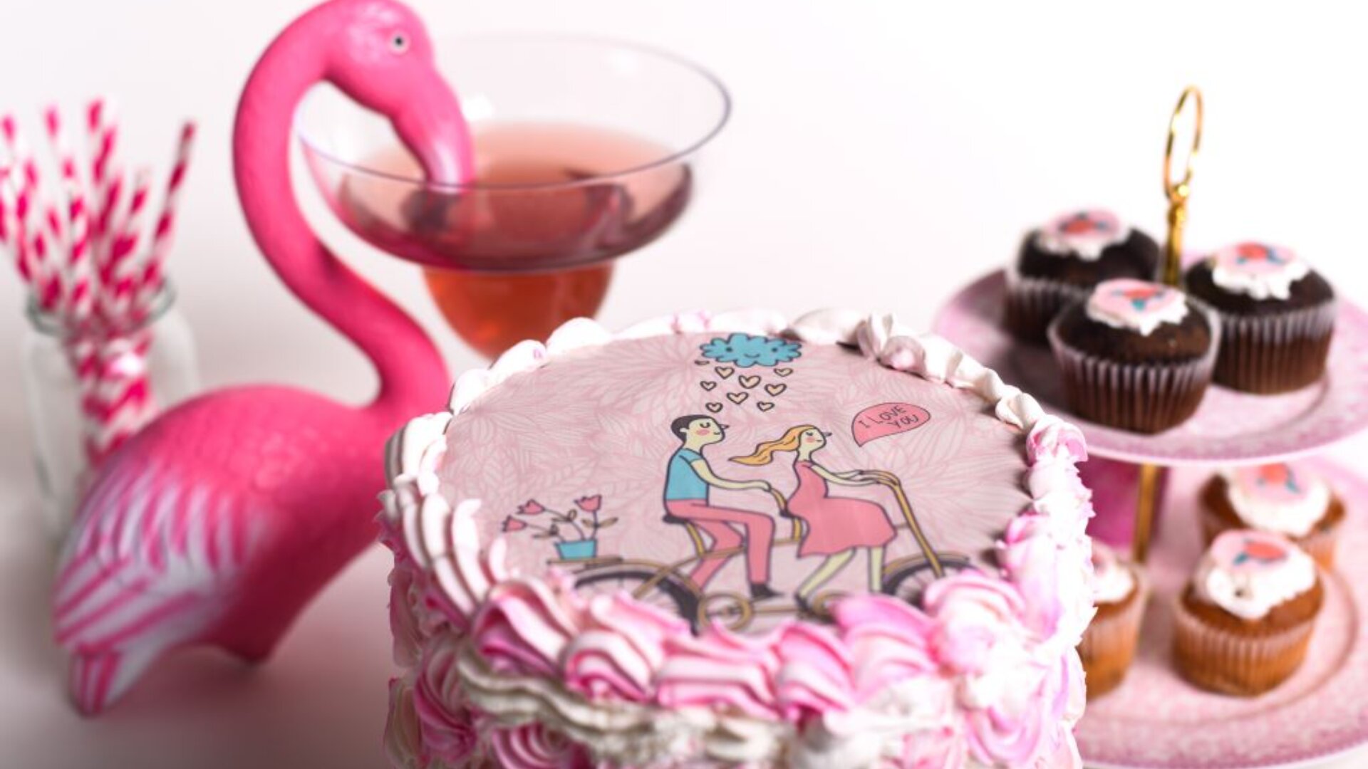A pink and white cake with a cartoon couple riding a bicycle, surrounded by pink flamingo decor, a cocktail, striped straws, and cupcakes topped with heart designs, creating a romantic celebration setting.