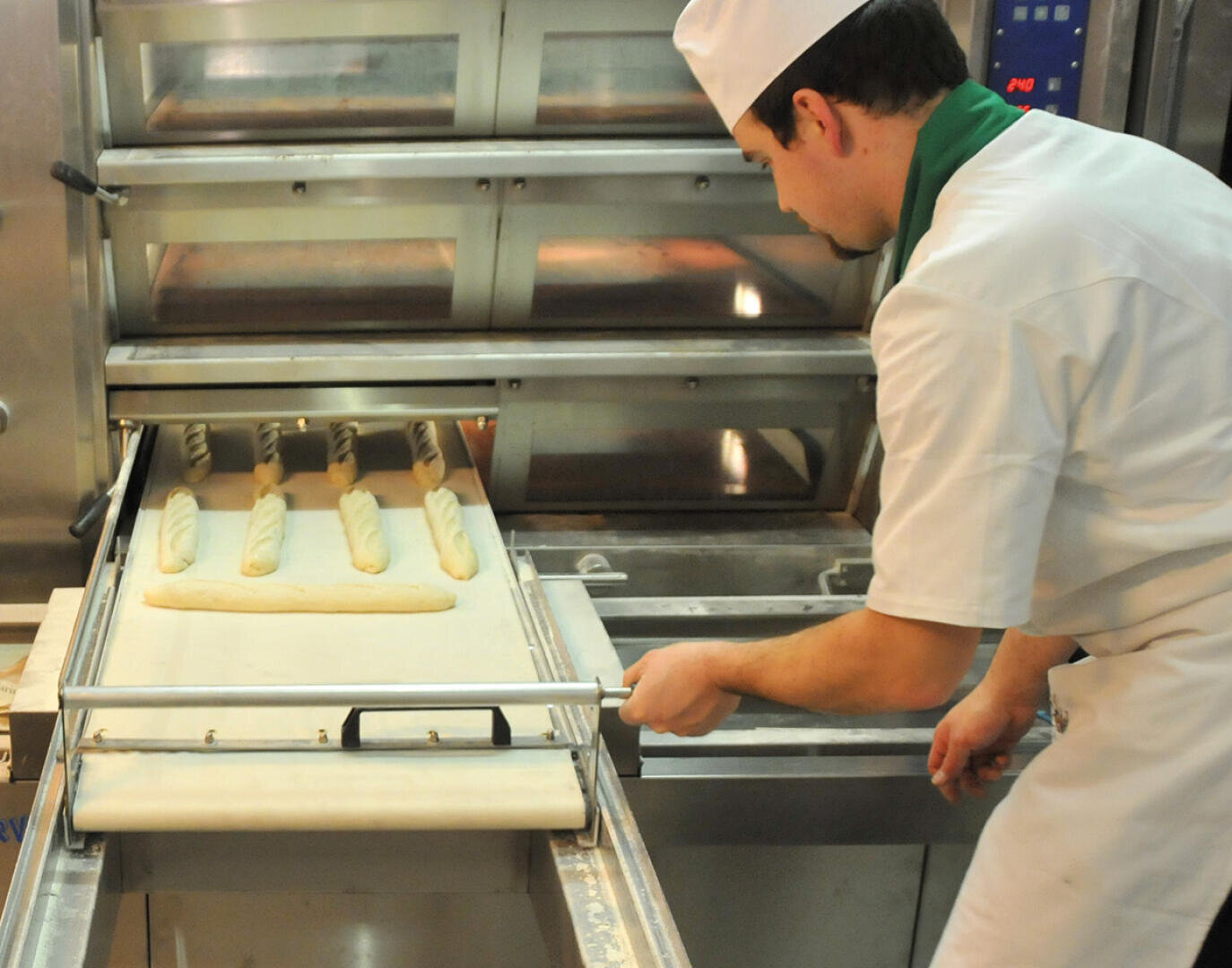 Ein Bäcker in weißer Uniform und mit Hut schiebt in einer Bäckereiküche Bleche mit rohem Baguette-Teig mit Hilfe eines Holzschälers in einen Industrieofen.