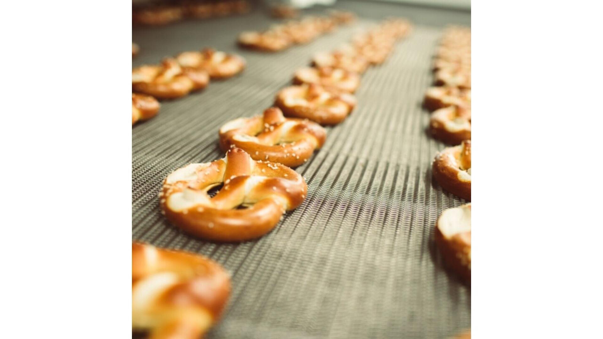 Rows of freshly baked pretzels are lined up on a conveyor belt, moving through what appears to be a bakery or food production facility. The pretzels are golden brown and sprinkled with coarse salt.