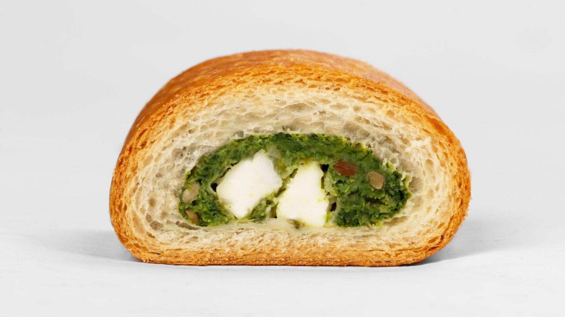A cross-section of a baked pastry with a golden-brown crust, showing a green spinach filling and cubes of white cheese inside, set against a plain light background.