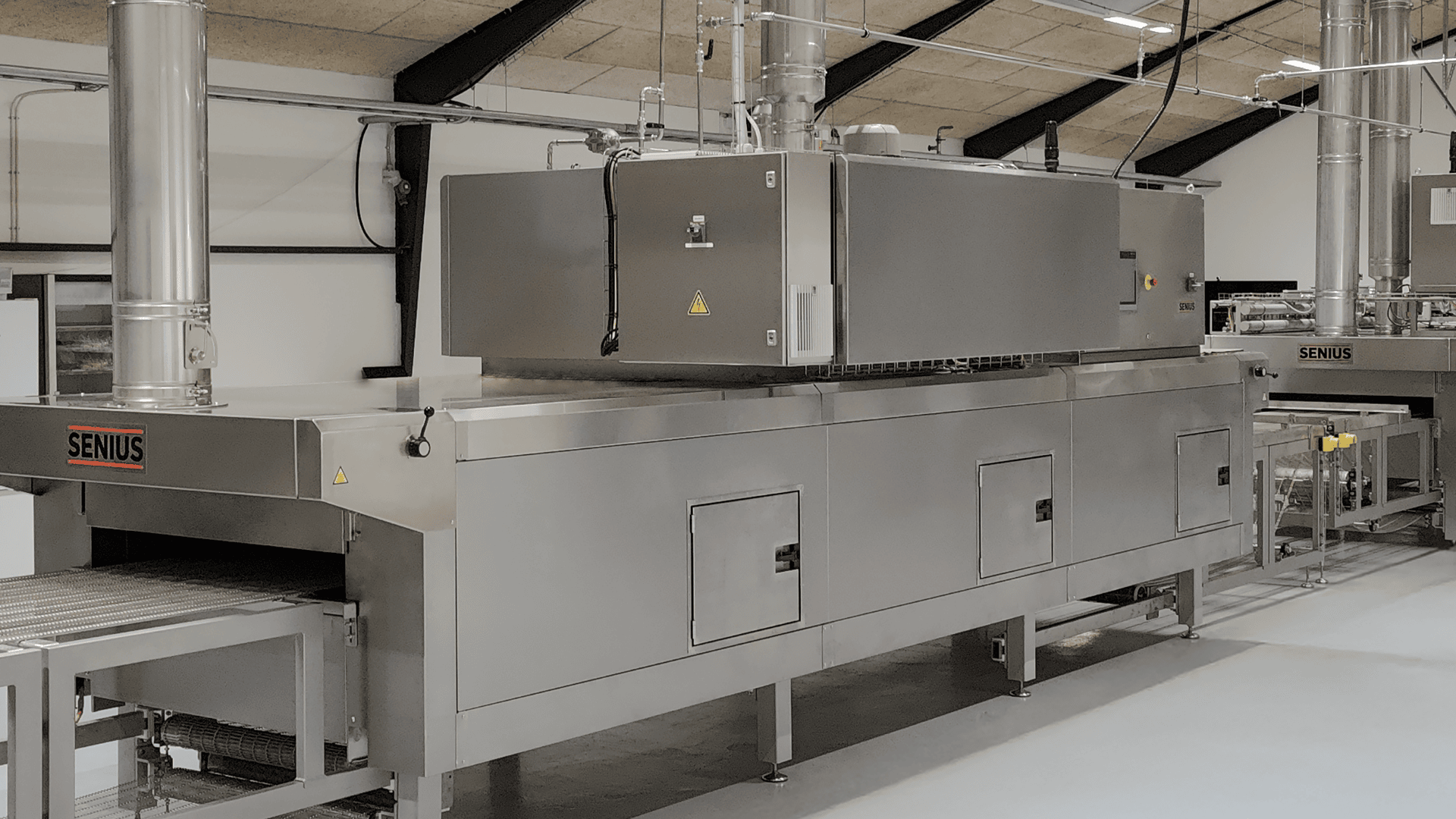 Großer Industrieofen aus rostfreiem Stahl mit Förderbändern und Lüftungsrohren, der für die gewerbliche Lebensmittelverarbeitung eingesetzt wird. Die Maschine befindet sich in einer sauberen, modernen Fabrikumgebung.