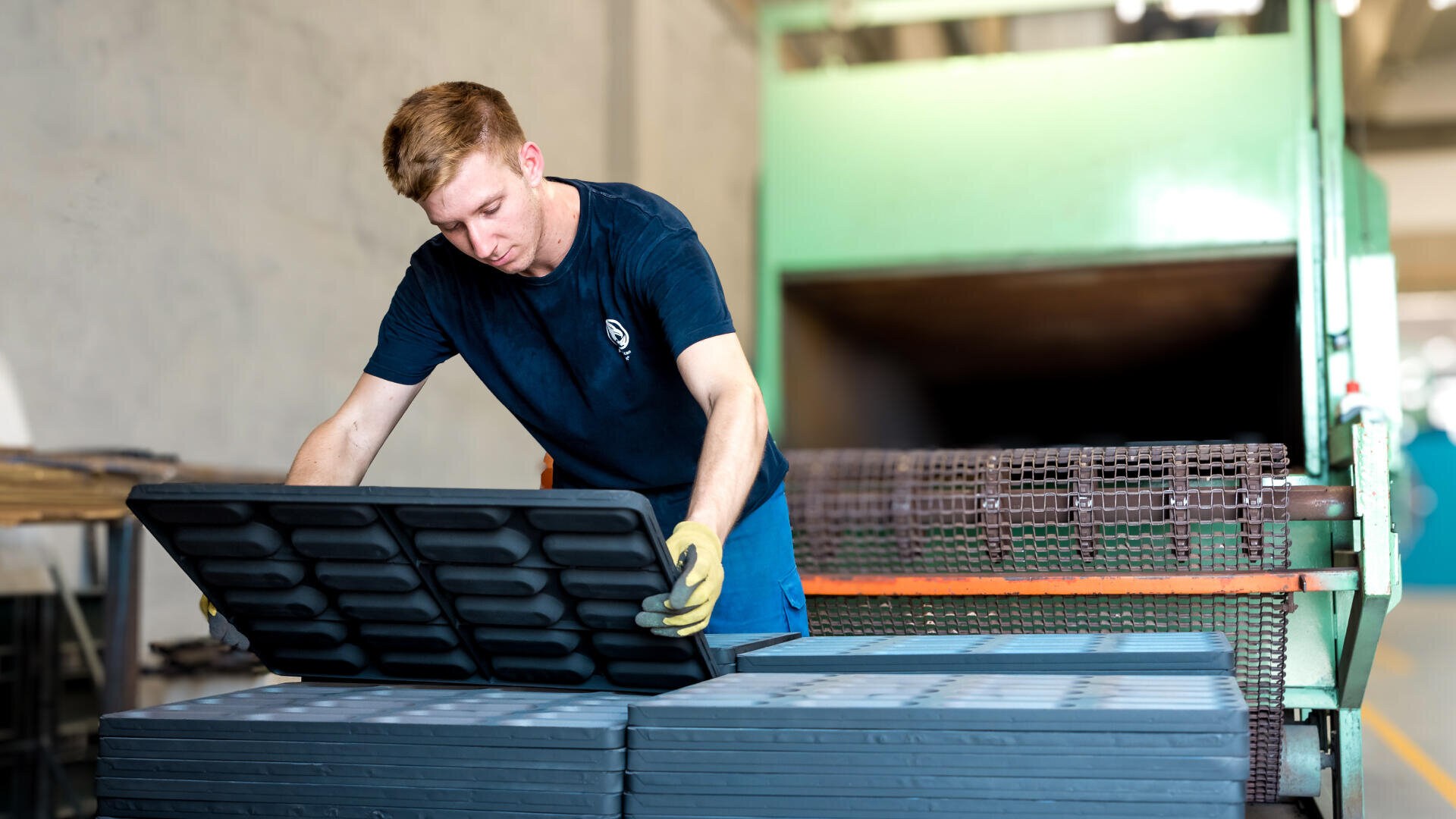 Ein Mann mit Handschuhen stapelt oder inspiziert große schwarze Plastikschalen in der Nähe von Industriemaschinen in einer Fabrik.