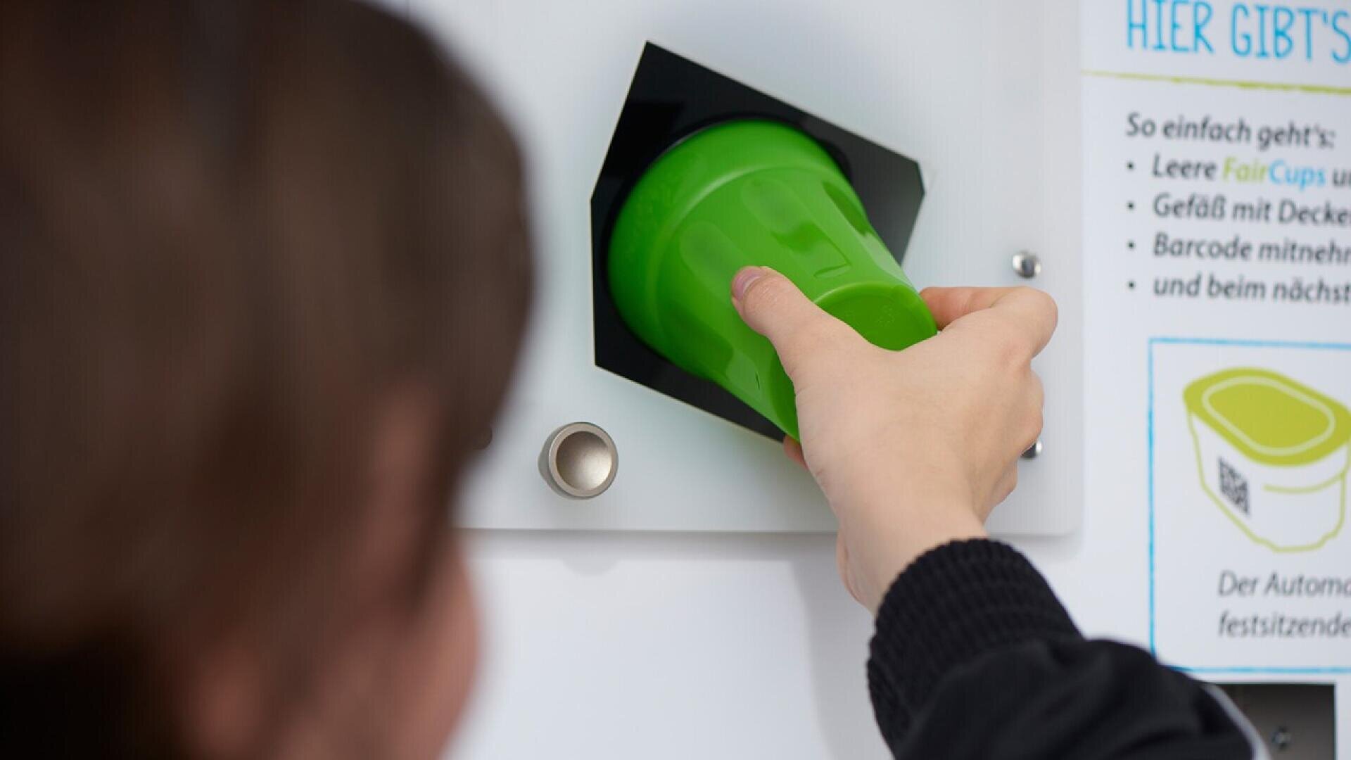 A person inserts a green reusable cup into a return slot of a vending or recycling machine, with instructions visible on the right side of the image.