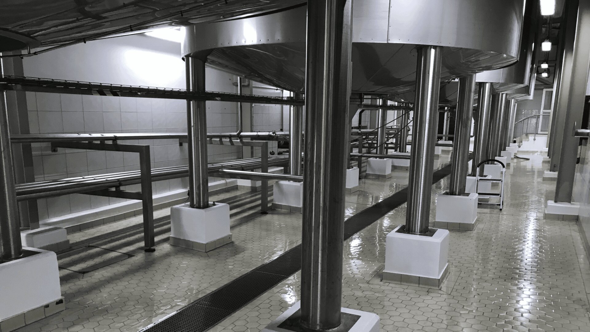 Industrial facility interior with large metal tanks elevated on steel columns, visible piping, tiled walls, and a hexagonal tile floor. The space is clean and well-lit, suggesting a food or chemical processing area.