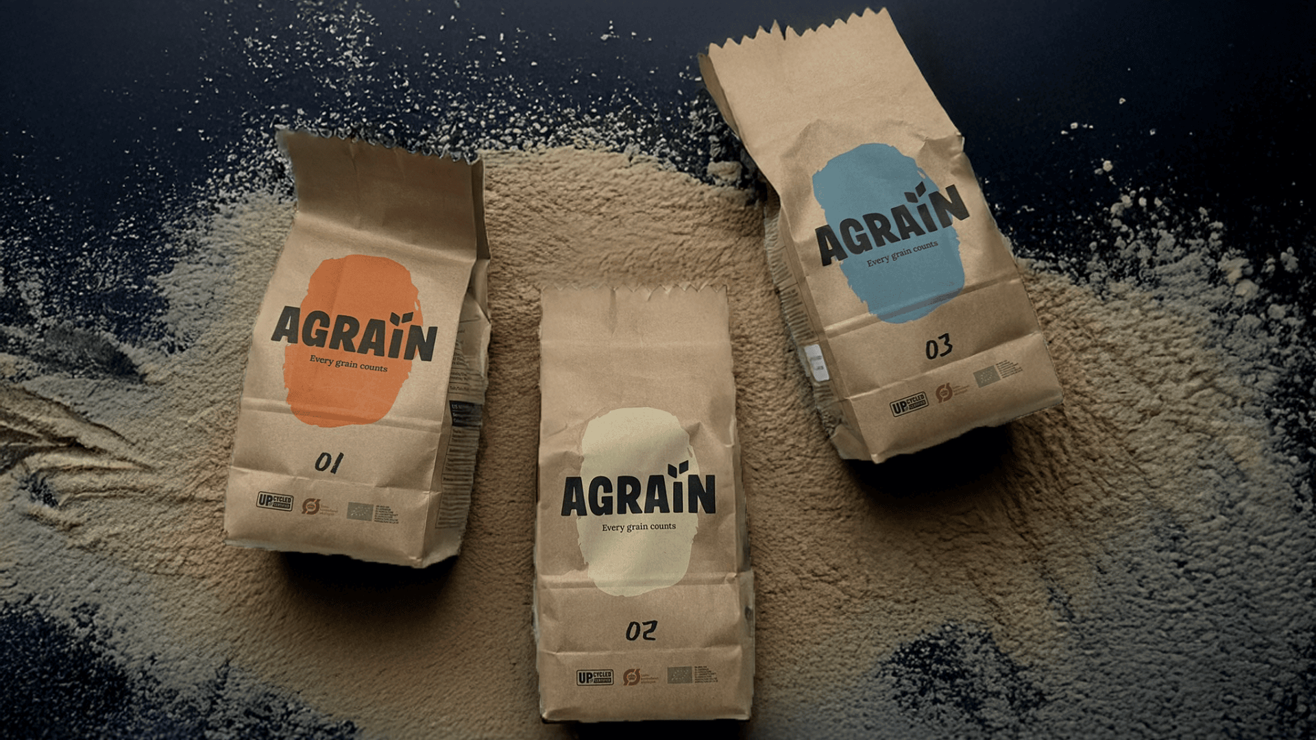 Three brown paper bags labeled AGRAIN with different colored labels (orange, blue, and teal) are placed on a dark surface dusted with flour or grain, arranged in a slightly angled, casual manner.