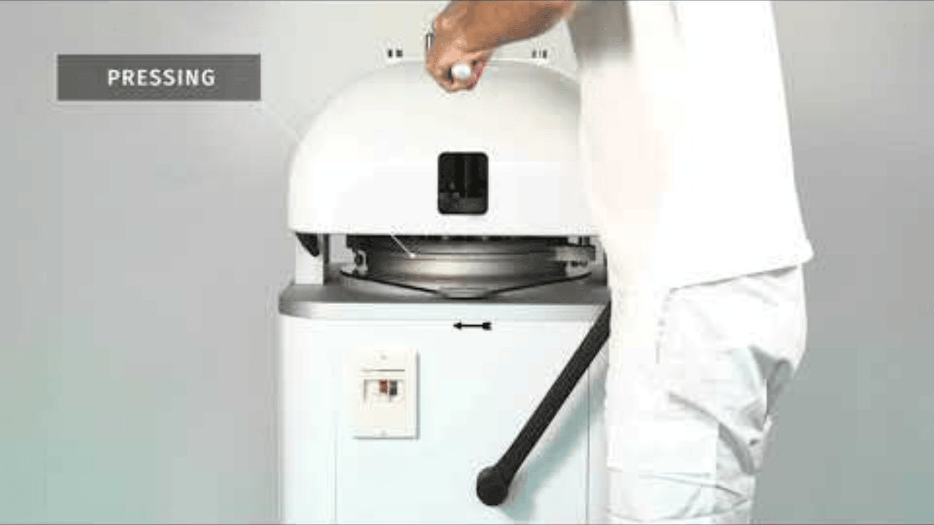 Eine Person bedient eine weiße Industriemaschine, indem sie auf einen Griff drückt. Das Wort PRESSING erscheint in einem grauen Kasten in der oberen linken Ecke. Die Maschine hat eine runde, kuppelförmige Oberseite und steht vor einem einfarbigen Hintergrund.