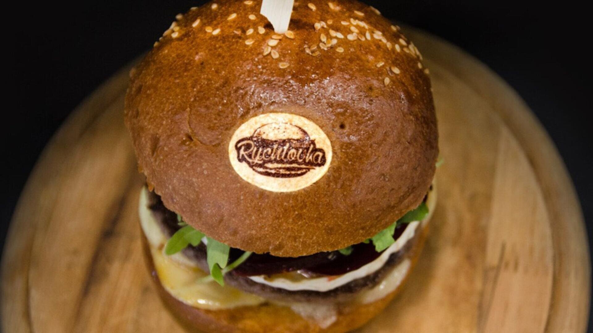Nahaufnahme eines Gourmet-Hamburgers auf einer hölzernen Unterlage mit einem glänzenden Sesambrötchen mit Logo, frischem Gemüse, Käse und einer dicken Rindfleischpastete, die mit einem Holzspieß befestigt ist.