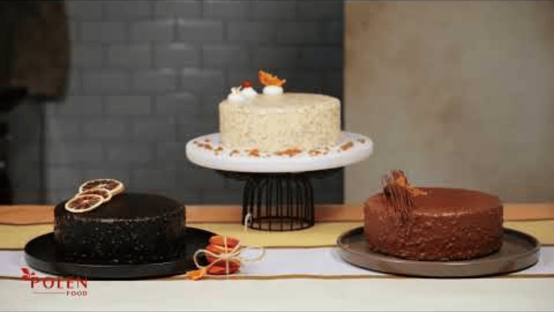 Three cakes are on display: a white cake with decoration on a stand in the center, a dark chocolate cake with dried orange slices on the left and a milk chocolate cake with chocolate decoration on the right.