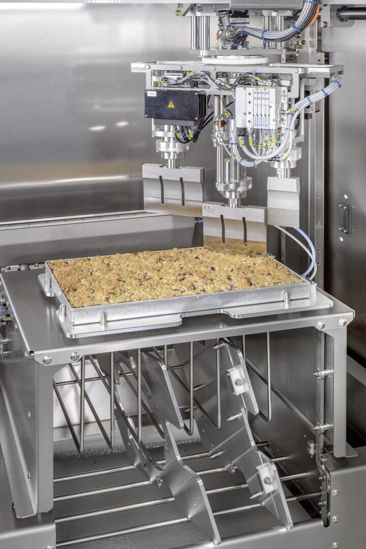 Nahaufnahme einer industriellen Lebensmittelverarbeitungsmaschine, die ein mit einer gebackenen Mischung gefülltes Backblech auf ein Metallgestell in einer Edelstahlumgebung abstellt.