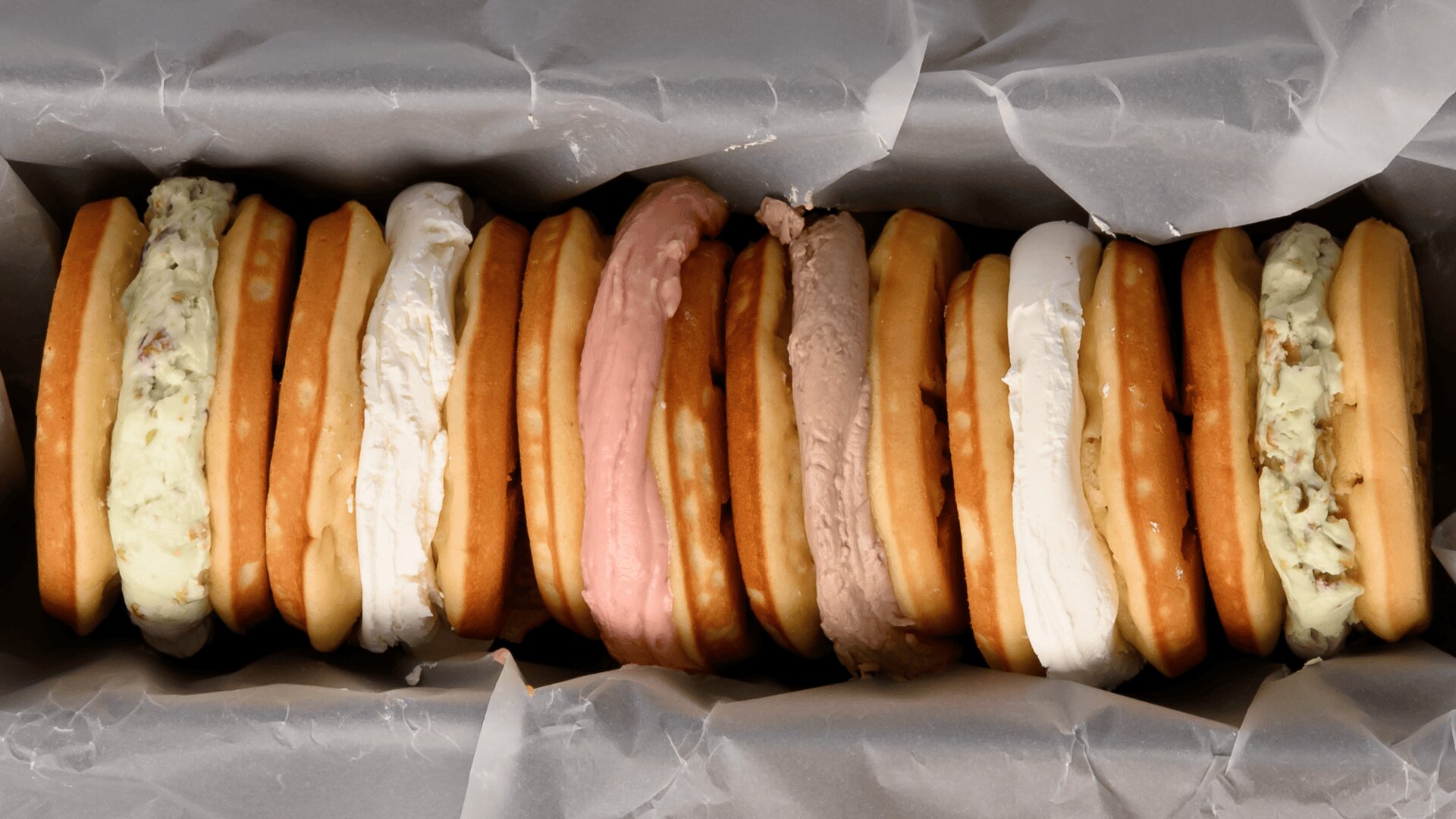 A row of round ice cream sandwiches with various flavors is lined up in a box, separated by wax paper. The ice cream fillings include shades of white, pink, brown, and green.