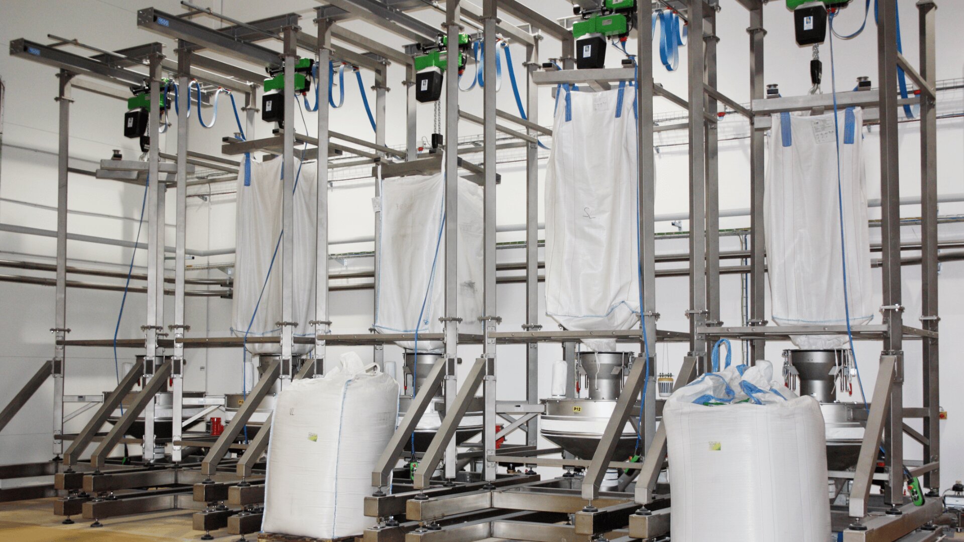 Five large white industrial bulk bags supported by a stainless steel frame, with machinery and pipes overhead, in a clean, modern factory setting. Two filled bags stand upright in front of the frame.