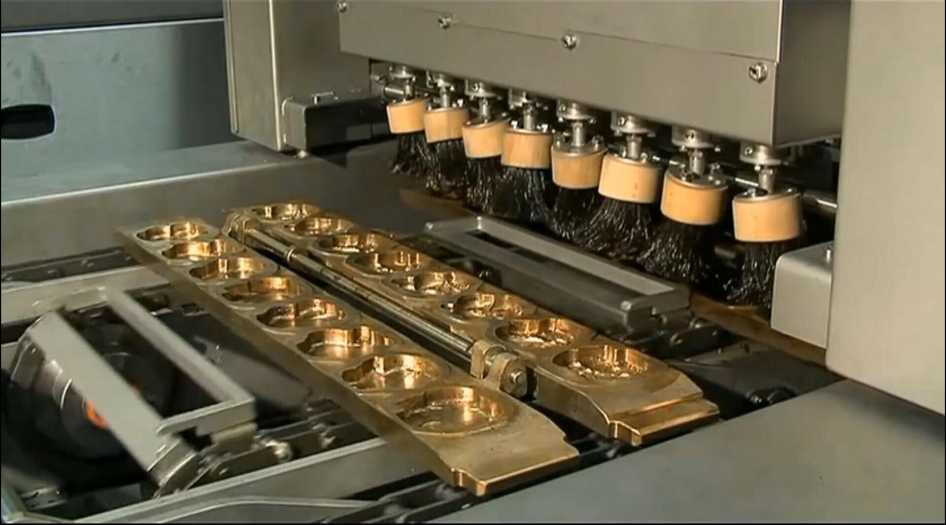 Eine Maschine mit mehreren rotierenden Bürsten poliert mehrere goldfarbene Metallteile, die in Reihen auf einem Förderband angeordnet sind.