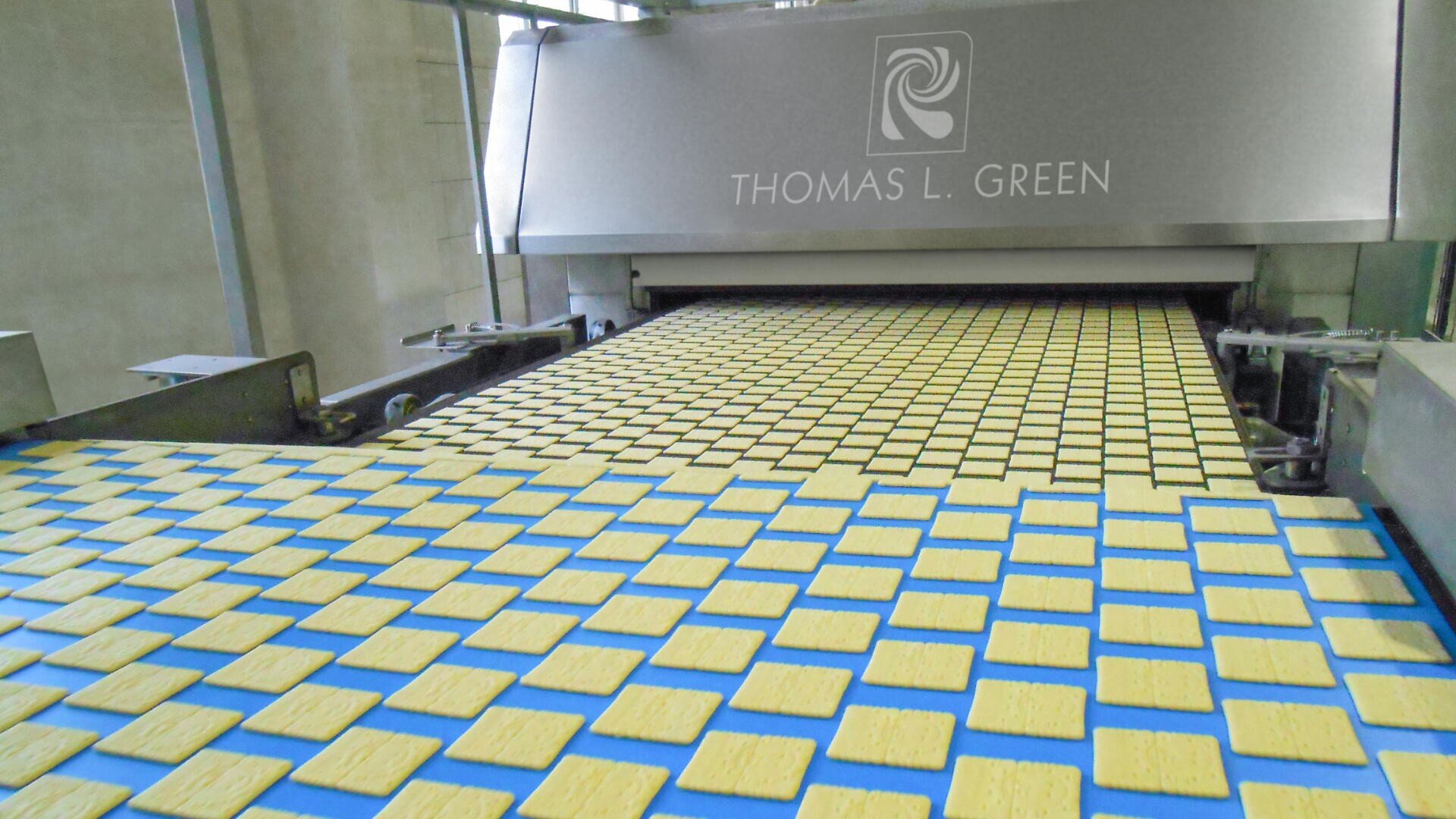 Rows of square crackers move along a conveyor belt into a large industrial baking machine labeled “THOMAS L. GREEN” in a food processing facility.