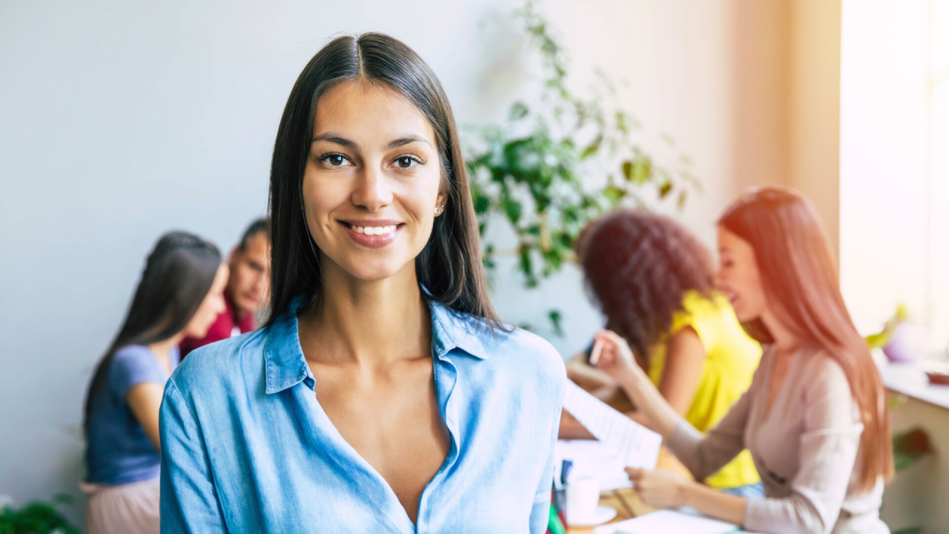 A woman in a blue shirt smiles at the camera, standing in an office with three colleagues working together at a table in the background. The atmosphere appears bright and collaborative.