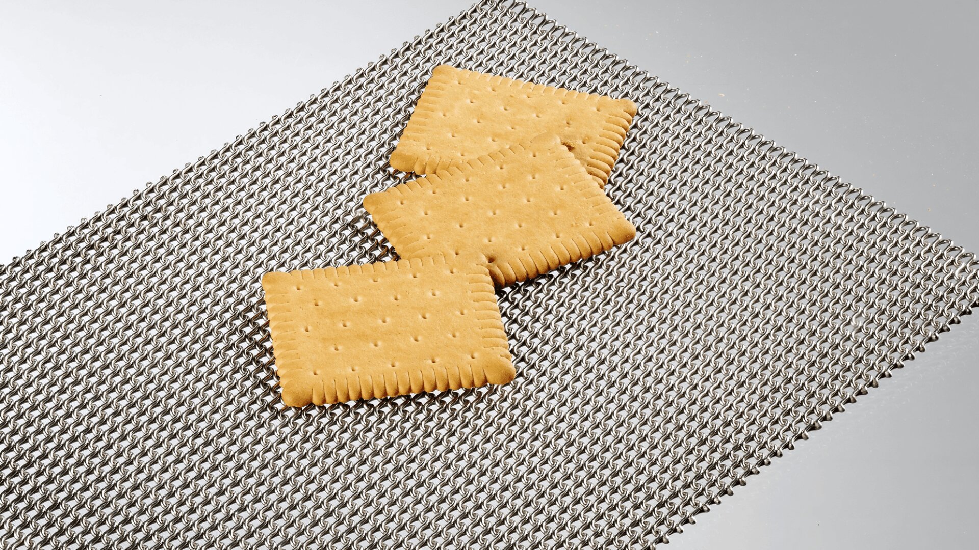 Drei rechteckige, goldbraune Cracker mit perforierter Oberfläche sind auf einer metallischen, strukturierten Matte vor einem schlichten, hellen Hintergrund angeordnet.