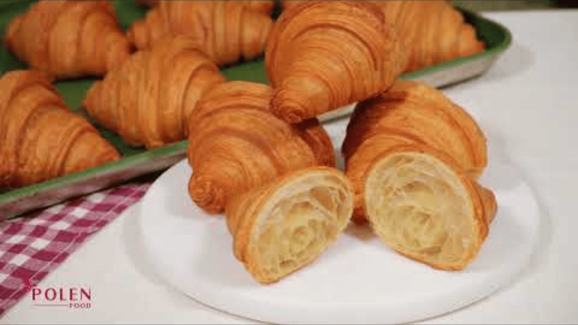 Several golden-brown croissants are displayed on a tray, with two croissants cut in half on a white plate, showing their flaky, layered interior. A red and white cloth is partially visible in the corner.