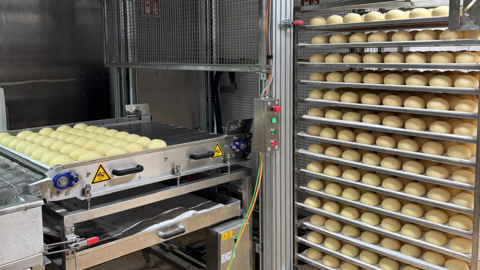 Reihen von Teigbällchen auf Metallblechen liegen neben einer Industriemaschine in einer Großbäckerei. Der Teig ist auf einem Förderband aufgereiht und die Bleche sind zum Backen vertikal gestapelt.