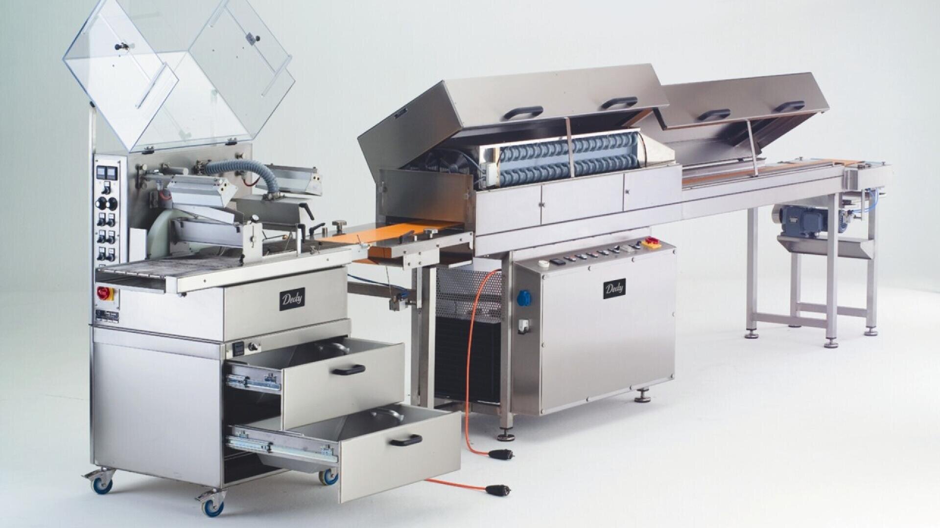 A large, stainless steel industrial food processing machine with conveyor belts, control panels, drawers, and open lids, designed for automated production in a commercial kitchen or factory setting.
