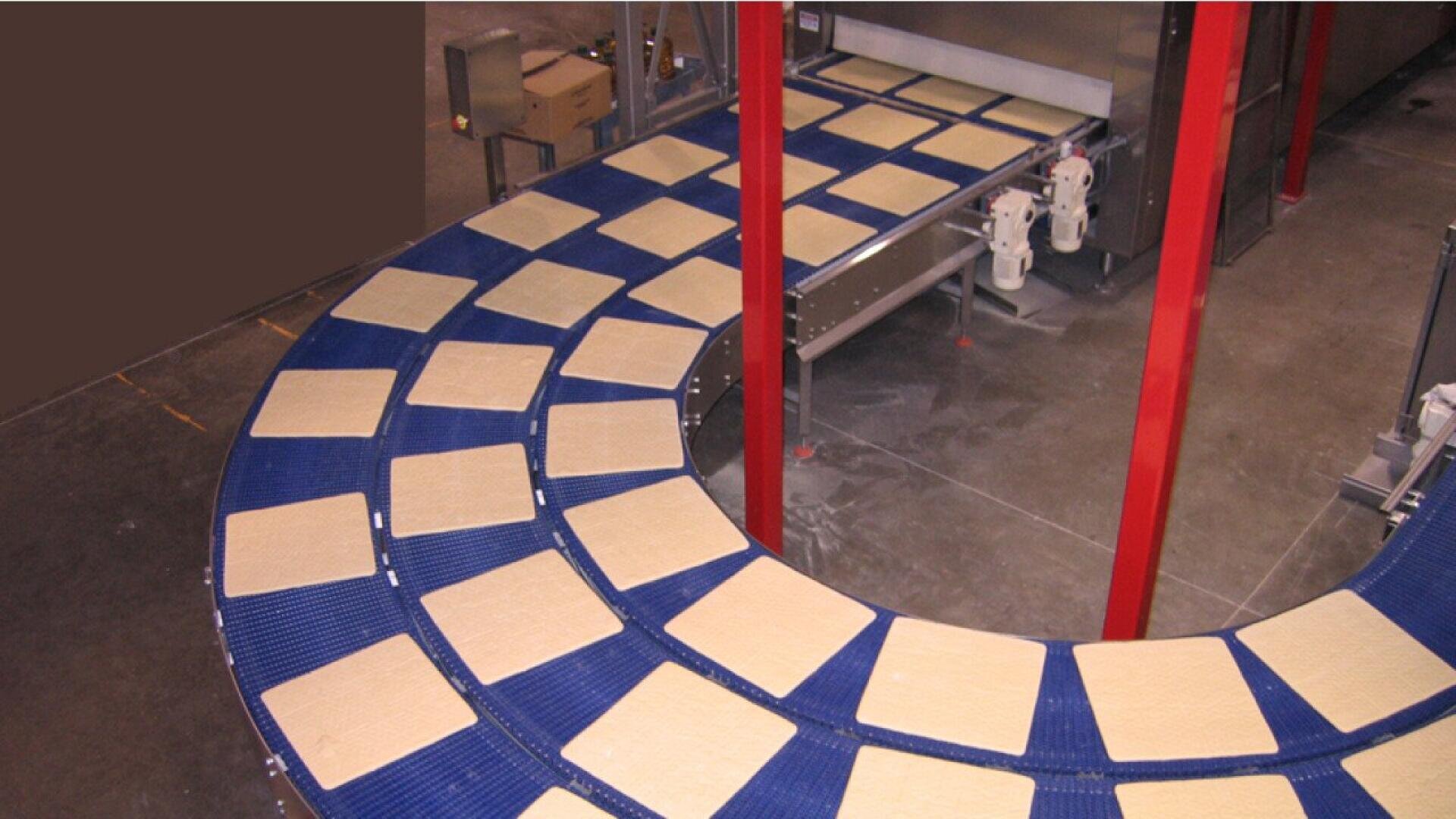 Rechteckige Teigblätter bewegen sich über ein gebogenes blaues Förderband in einer Lebensmittelverarbeitungsanlage, im Hintergrund sind Metallstrukturen und Maschinen zu sehen.