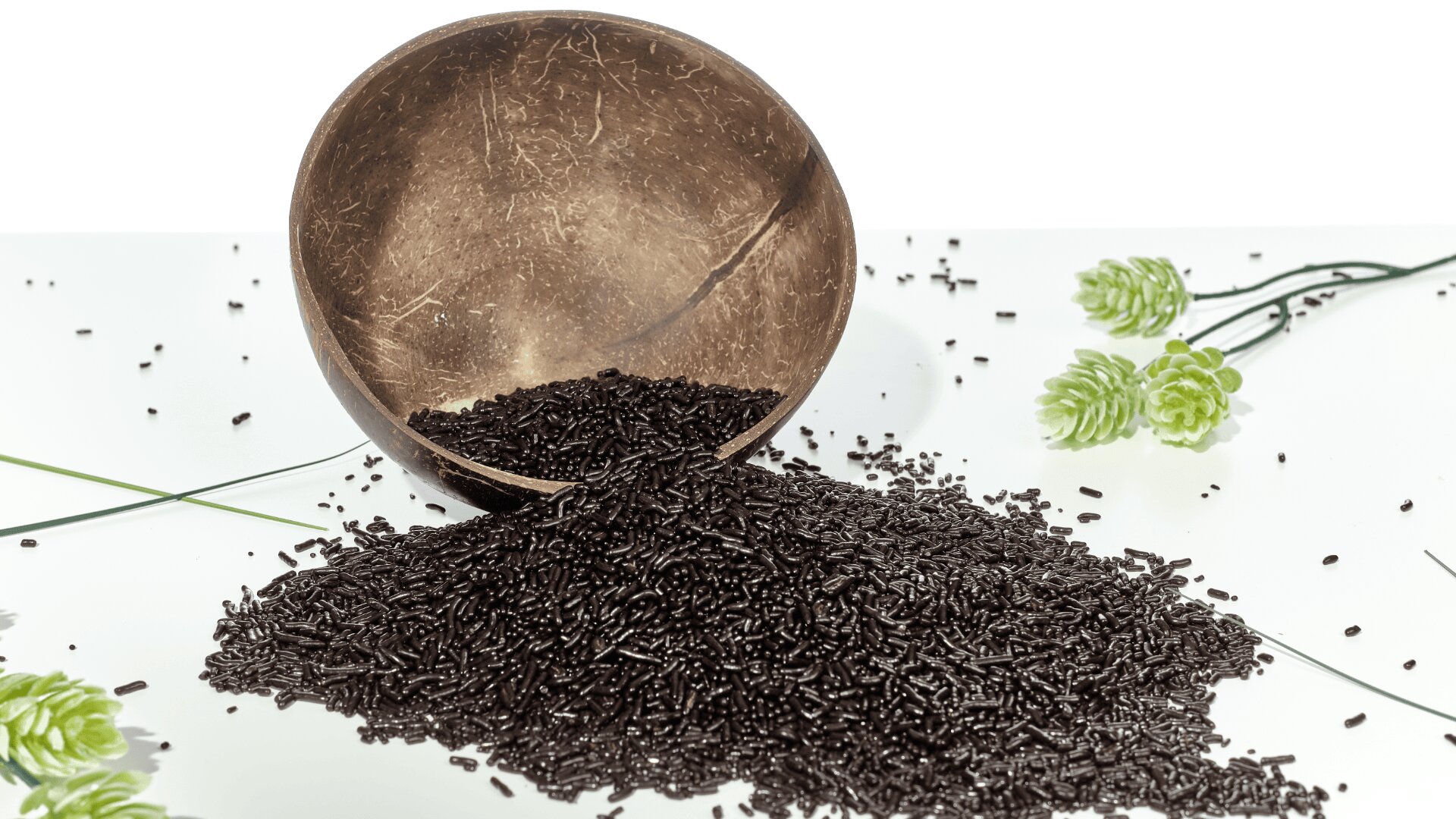 A coconut bowl tipped over, spilling black sprinkles onto a white surface. Green decorative flowers and thin green stems are arranged around the bowl and sprinkles.