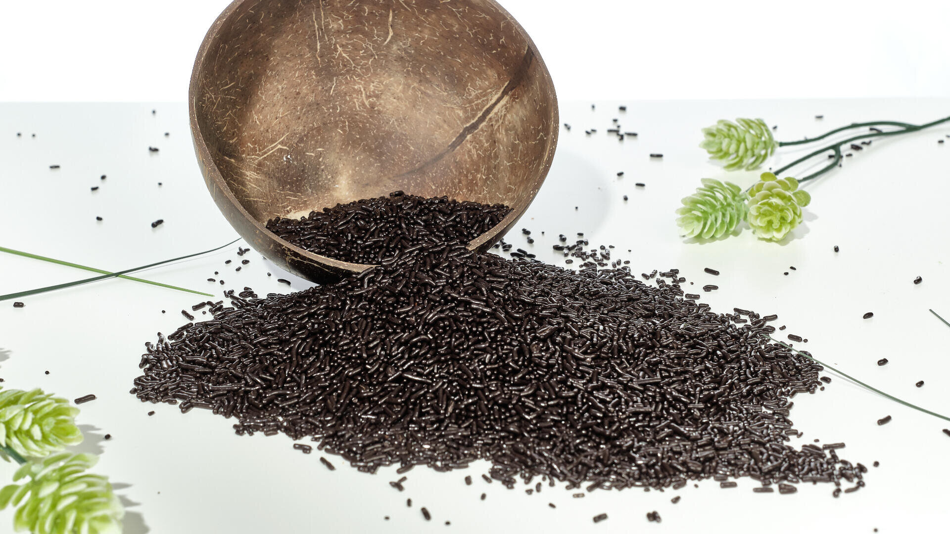 A coconut shell bowl tipped over, spilling a pile of dark chocolate sprinkles onto a white surface, with green artificial flowers scattered around.