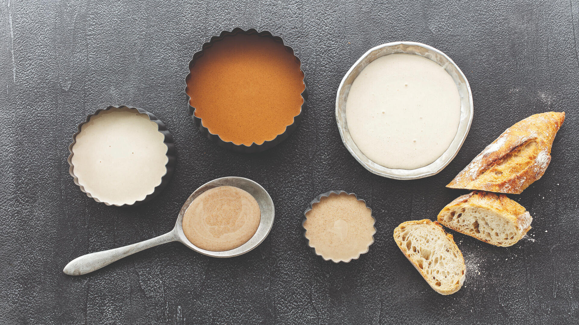 Mehrere kleine Schälchen und ein Löffel, gefüllt mit verschiedenen Arten von flüssigen Sauerteig-Startern, sind auf einer dunklen Fläche neben einem Laib und Scheiben von rustikalem Brot angeordnet.